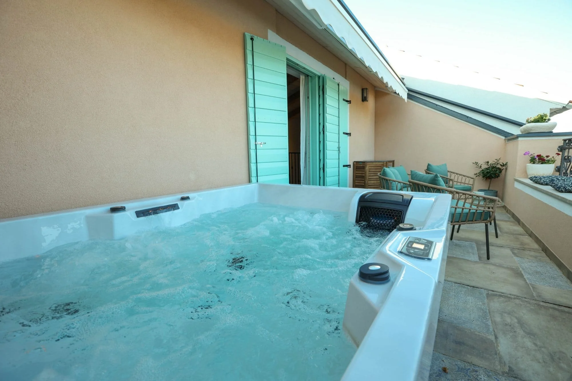 Outdoor balcony with a hot tub, wicker seating with cushions, potted plants, and an open door with turquoise shutters leading inside.