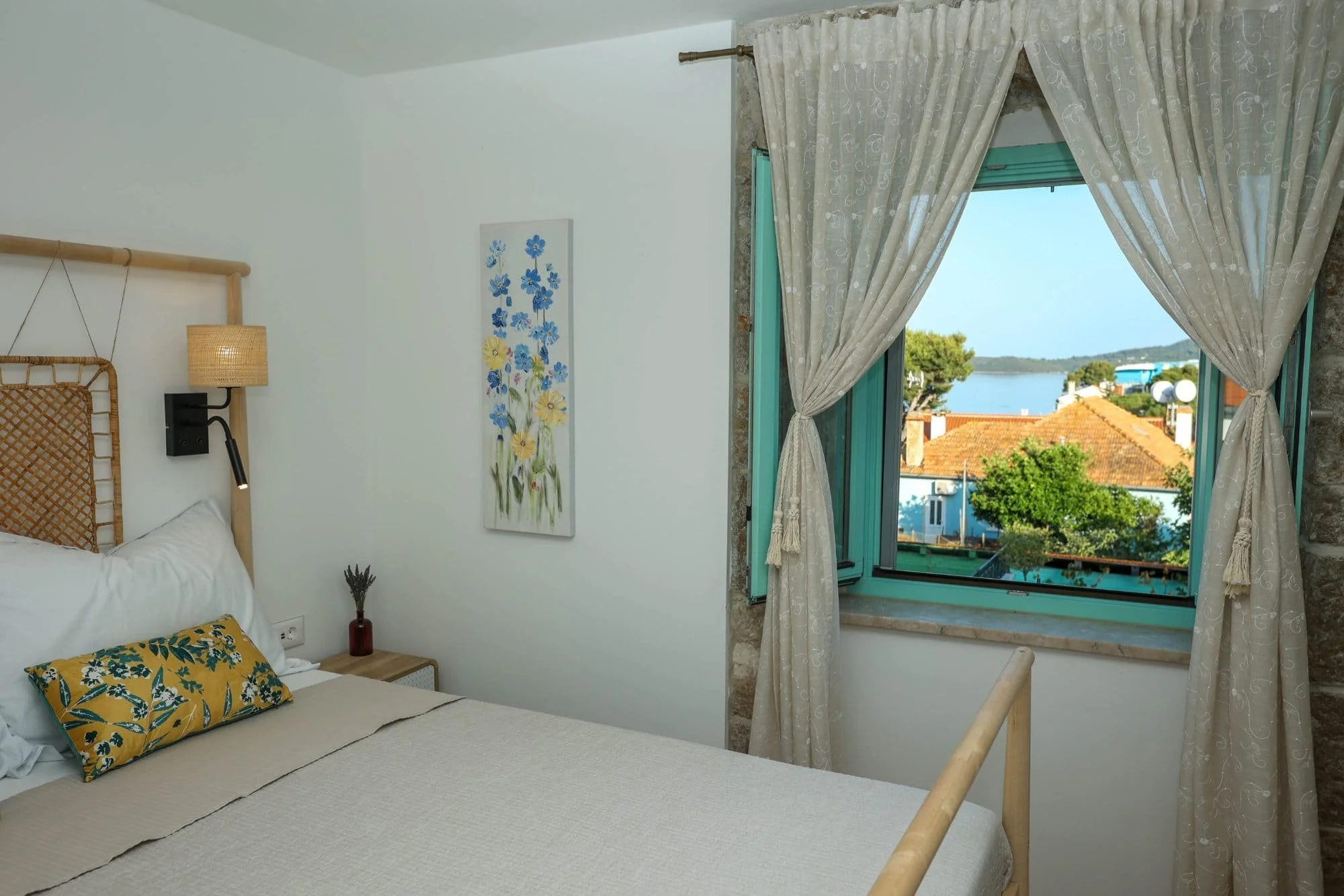 A bedroom with a white bed, floral pillow, wall painting, wooden nightstand, red vase with dried lavender, opened window with pale curtains, view of rooftops, trees, and water outside.