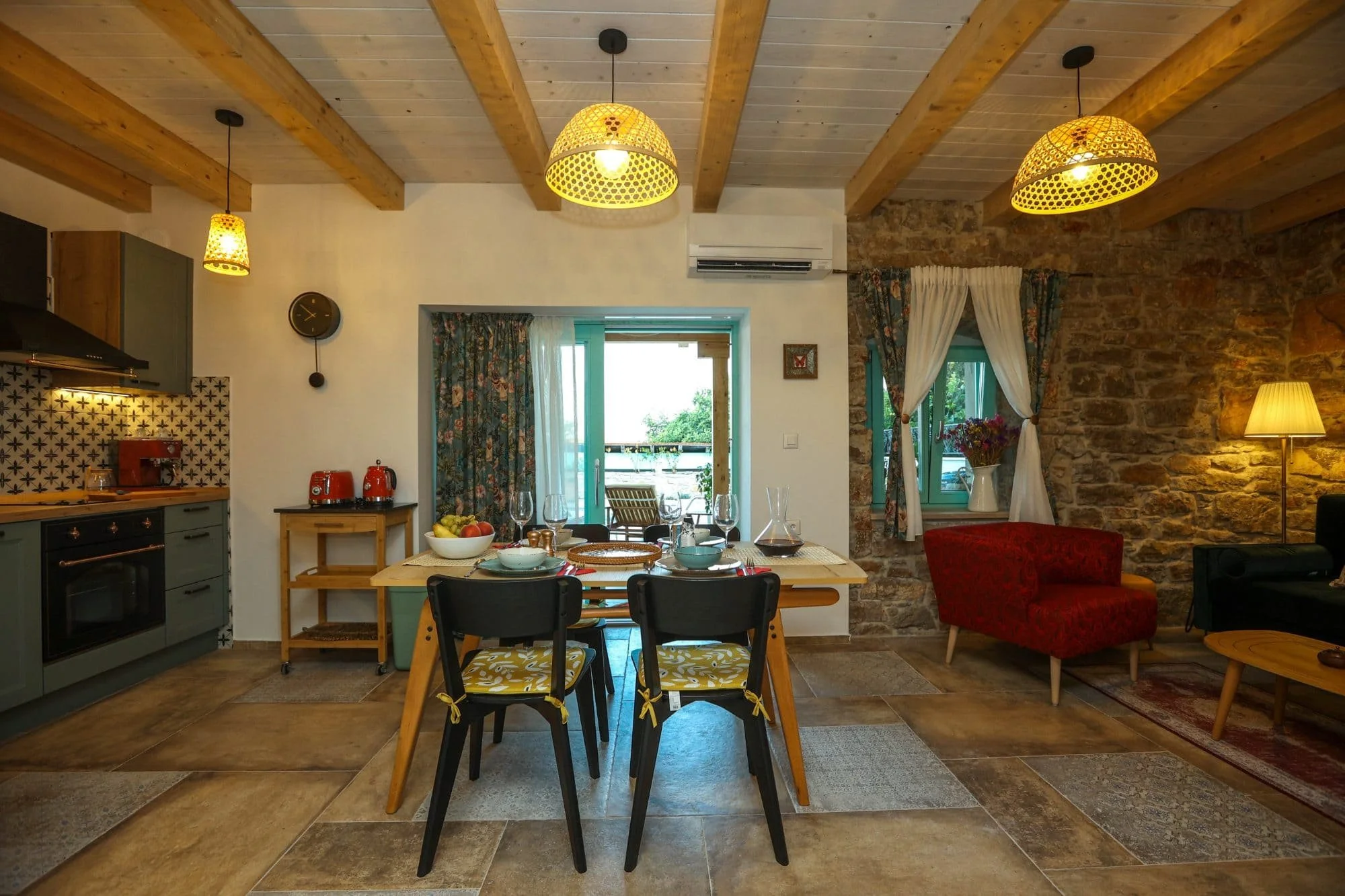 Interior of a cozy kitchen and dining area with rustic and modern decor. Wooden ceiling beams, yellow pendant lights, a stone wall, and large windows with floral curtains.