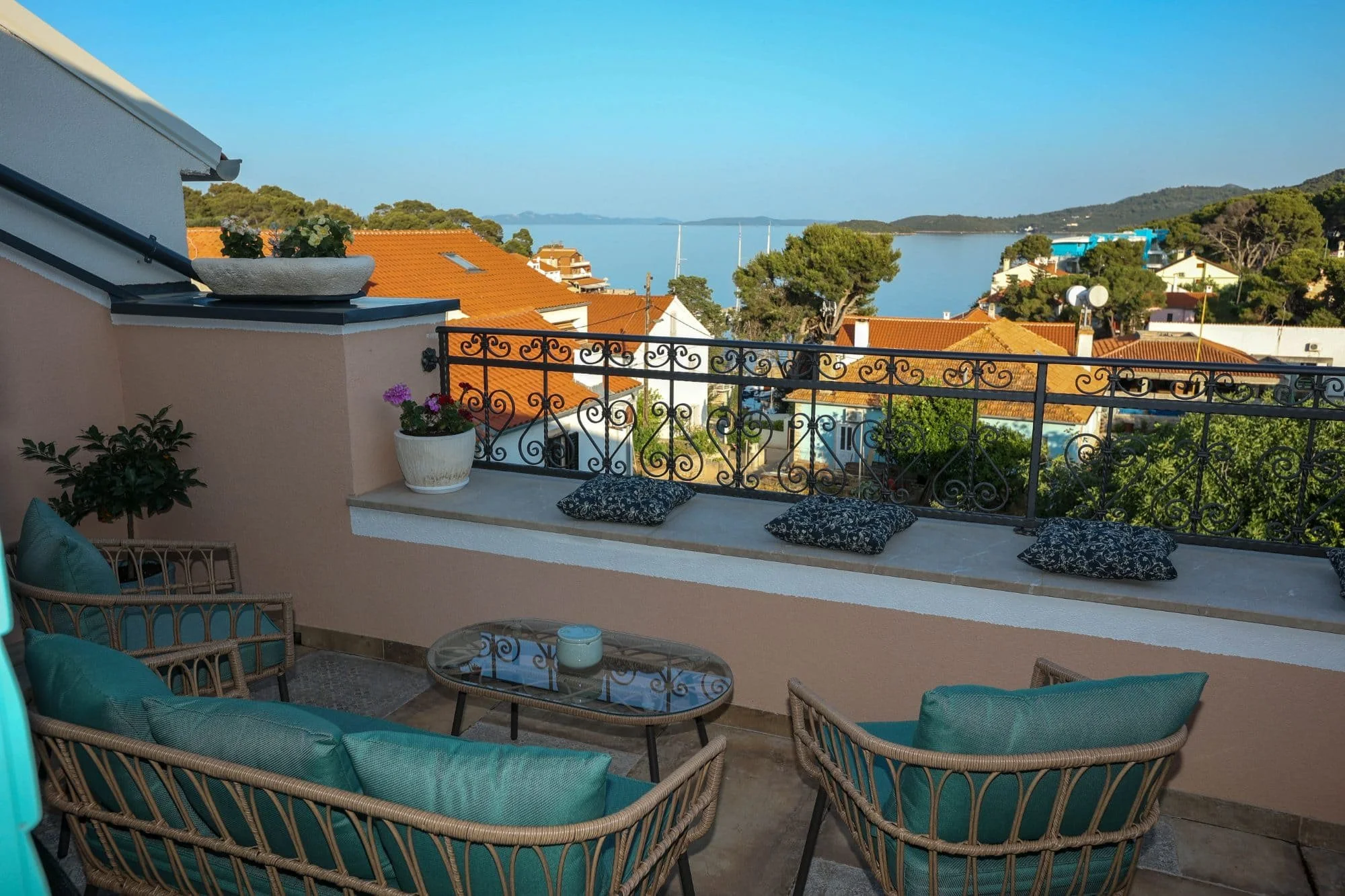 A balcony with wicker furniture, teal cushions, and black and white patterned pillows overlooking a neighborhood with orange-tiled roofs, trees, and a body of water in the distance.