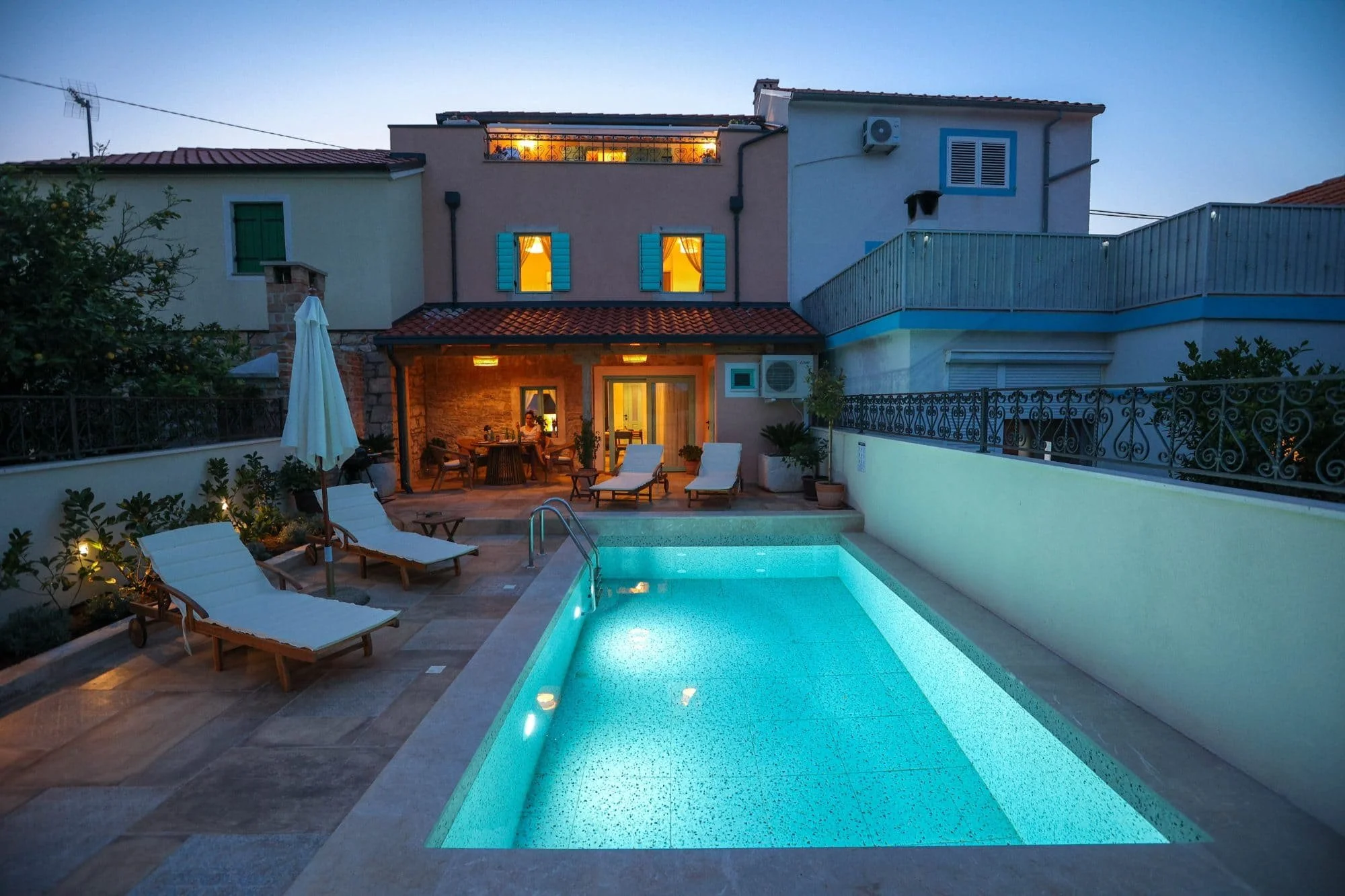Two-story house with outdoor pool, lounge chairs, and patio at dusk. Lights on inside, blue window shutters, rooftop terrace, and potted plants visible.