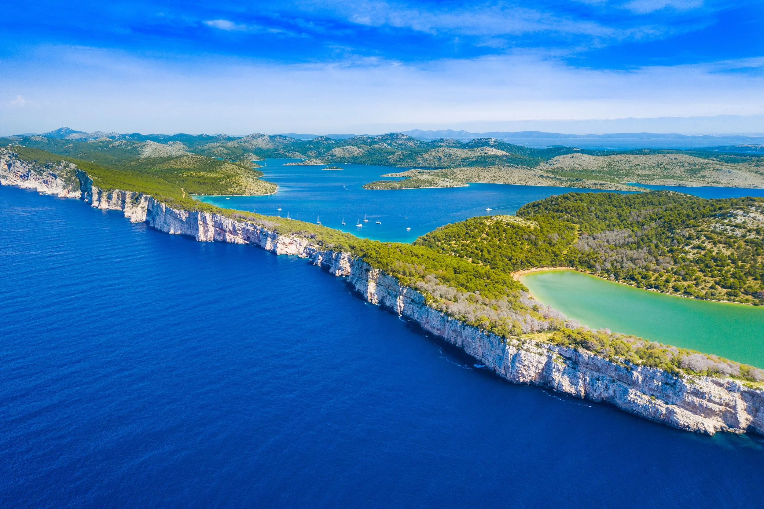 Aerial view of a coastal area with white cliffs, blue ocean, green forested hills, and a small lake with a boat and sailboats.