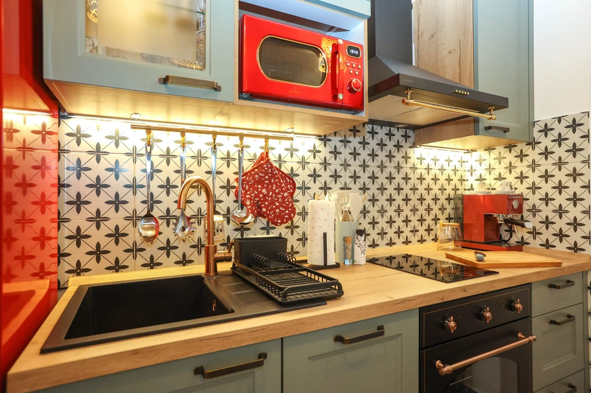 A modern kitchen with light blue cabinets, a wooden countertop, black sink, and a black stove. On the backsplash, there are black and white floral-patterned tiles. A red microwave, a roll of paper towels, utensils, and a red espresso machine are on t
