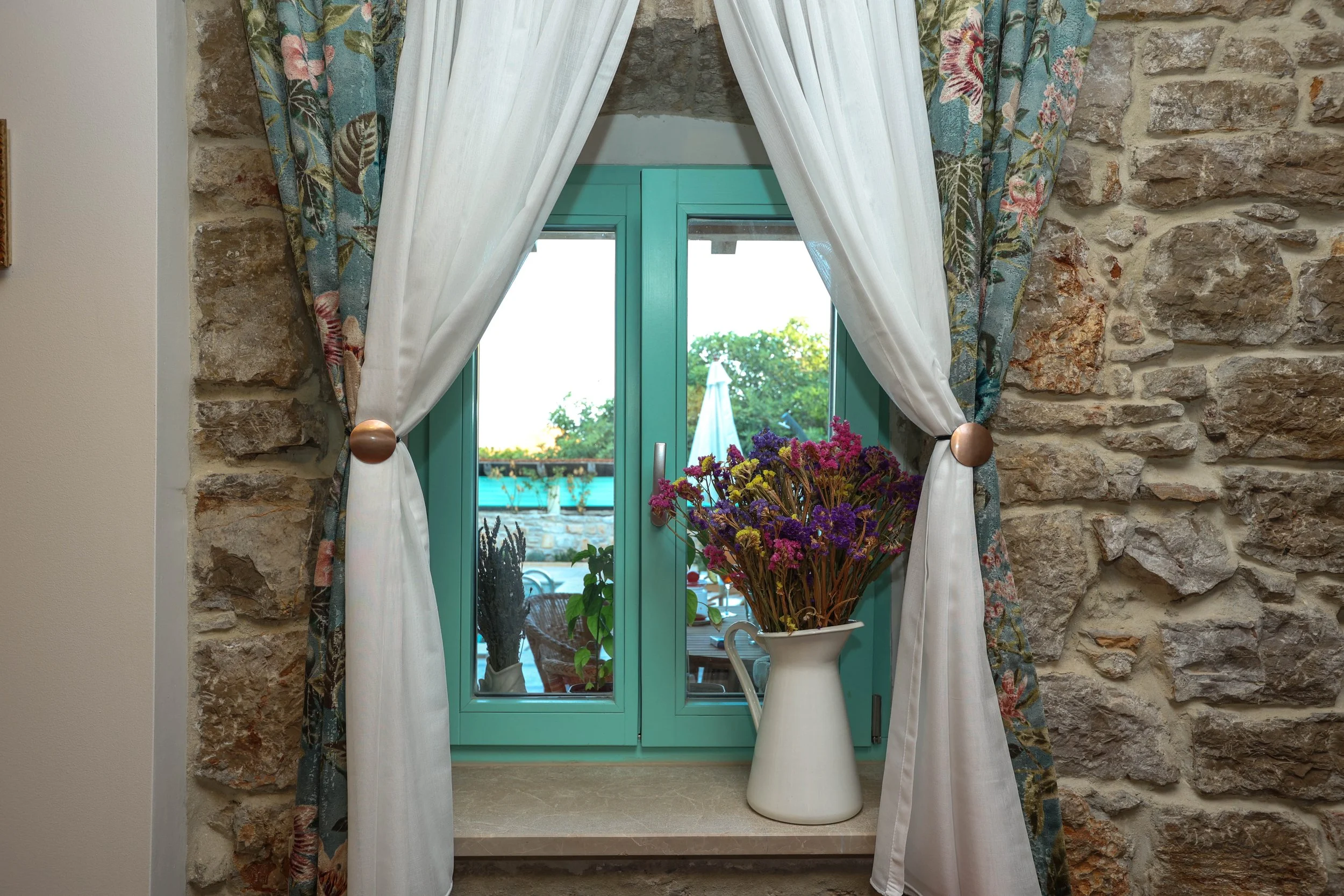 A window with turquoise frame surrounded by floral curtains, with a vase of colorful flowers on the windowsill and outside a patio with an umbrella and greenery.