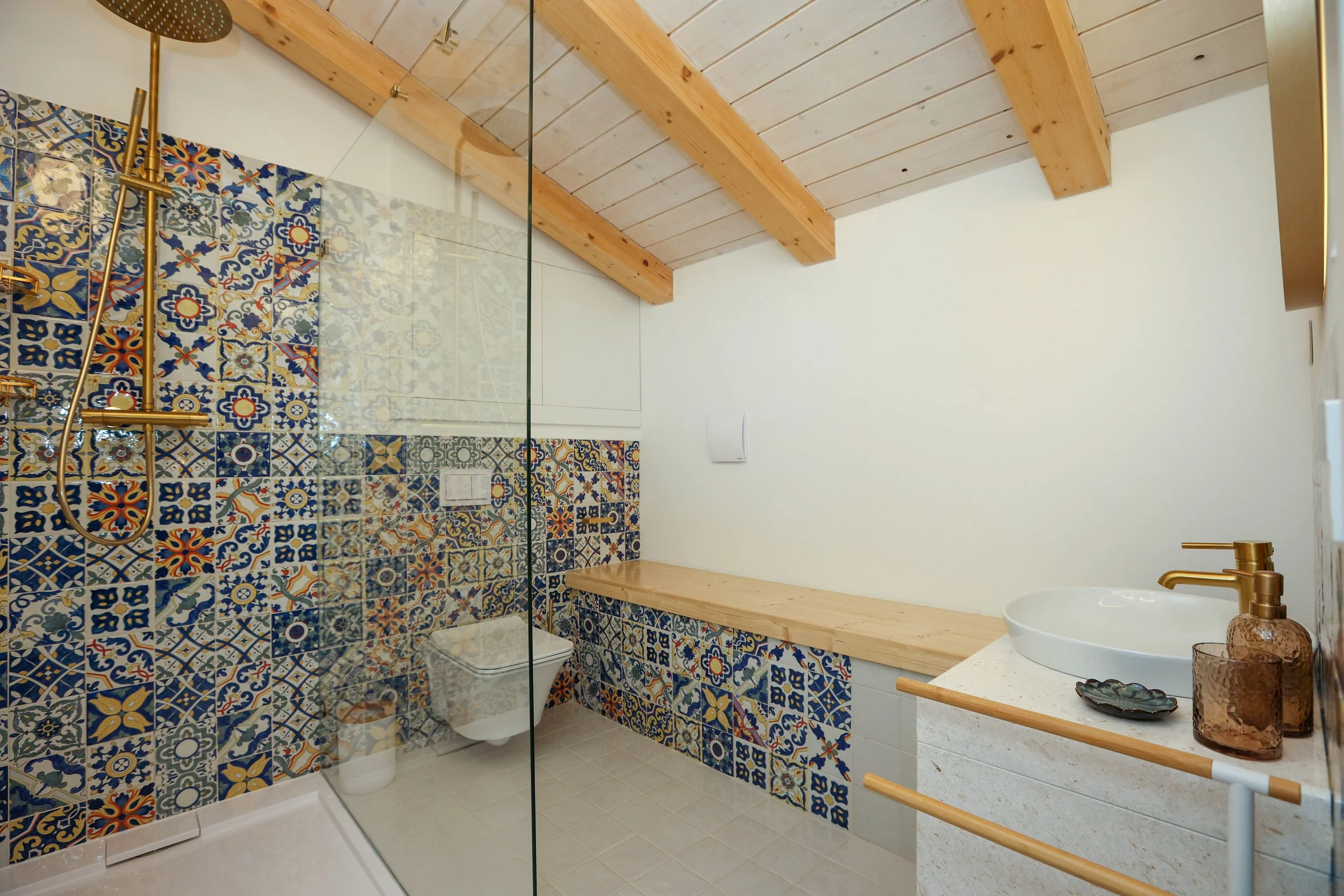 A modern bathroom featuring a walk-in shower with colorful patterned tiles, a white toilet, and a white vanity with a round vessel sink. The space has a wooden ceiling with exposed beams and a glass partition separating the shower area. There are decorative accessories on the vanity, including a soap dispenser, and a dark tray. The overall design combines bold tile patterns with natural wood elements.
