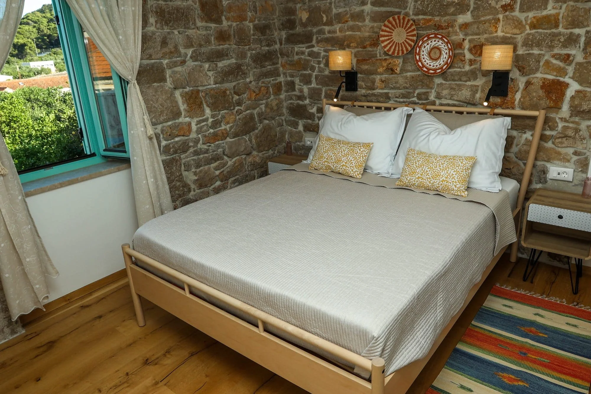 A cozy bedroom with a stone brick wall behind a wooden bedframe. The bed has white sheets and two yellow patterned pillows, flanked by two bedside tables with table lamps. There is a window with cream-colored curtains partially open, showing trees an