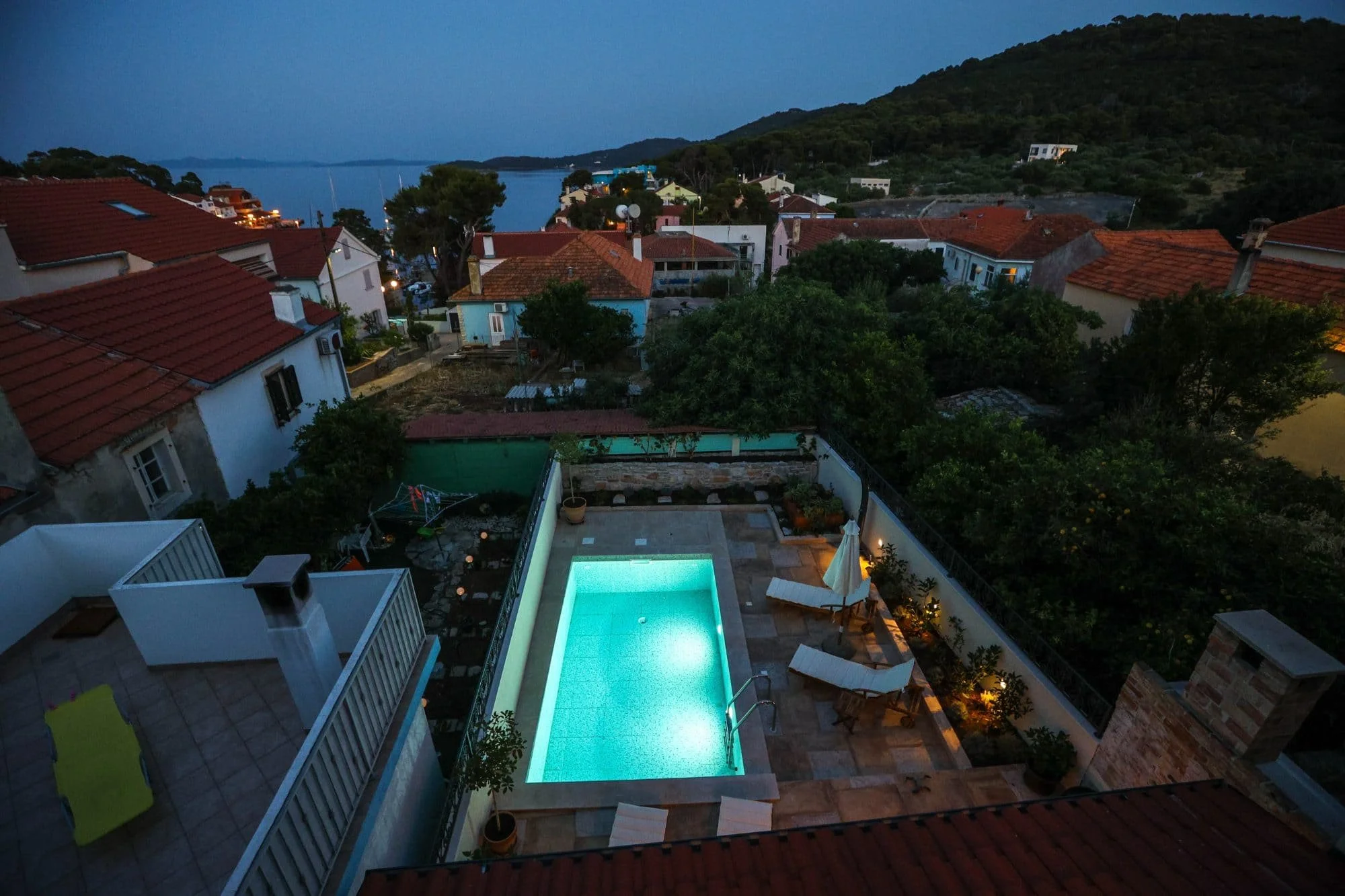 A nighttime view of a backyard with a lit swimming pool, surrounded by patio furniture, umbrellas, and lush greenery. In the background, rooftops of houses and hills are visible near a body of water under a dark sky.