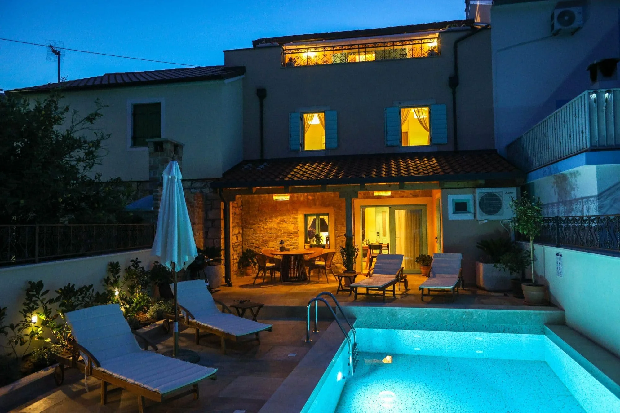 A house with lit windows at night, featuring a balcony and a patio area with a table, chairs, and poolside lounge chairs illuminated by pool lights.