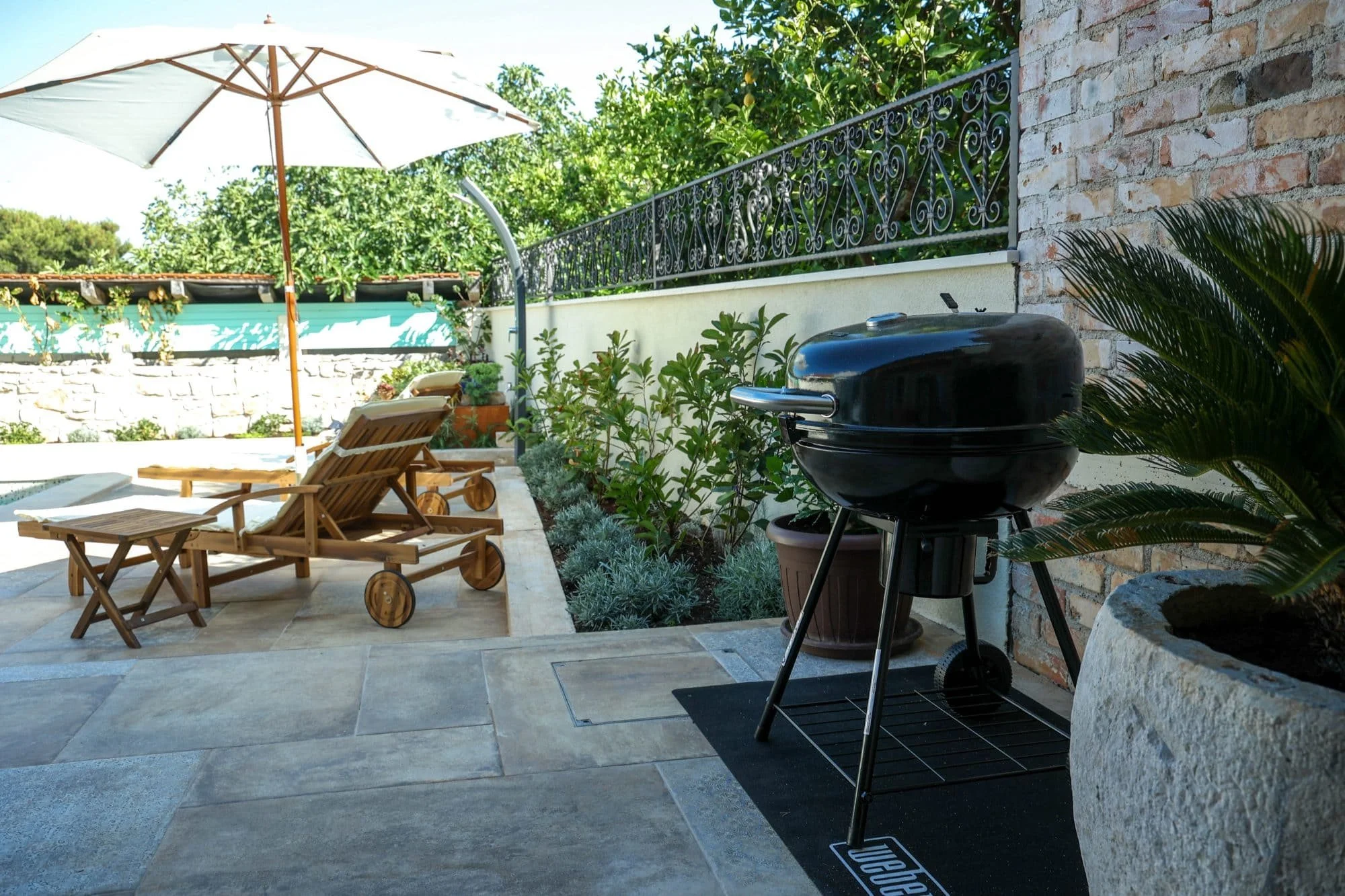 Outdoor patio with two wooden lounge chairs, a small table, a white patio umbrella, a black charcoal grill, and potted plants along a brick wall, with trees and greenery in the background.