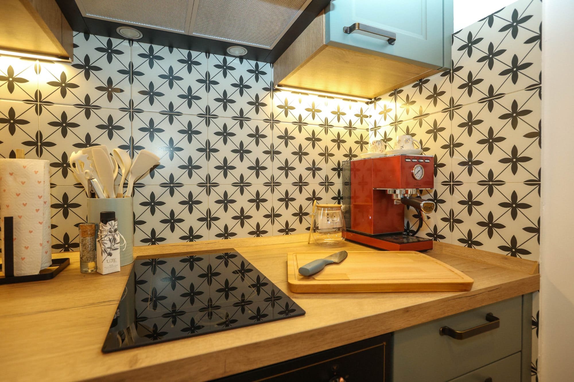 A kitchen countertop with a built-in black Cooktop, a red espresso machine, a glass jar with a white lid, a wooden cutting board, a white utensil holder with utensils, and a roll of paper towels. The backsplash has black and white floral-patterned ti