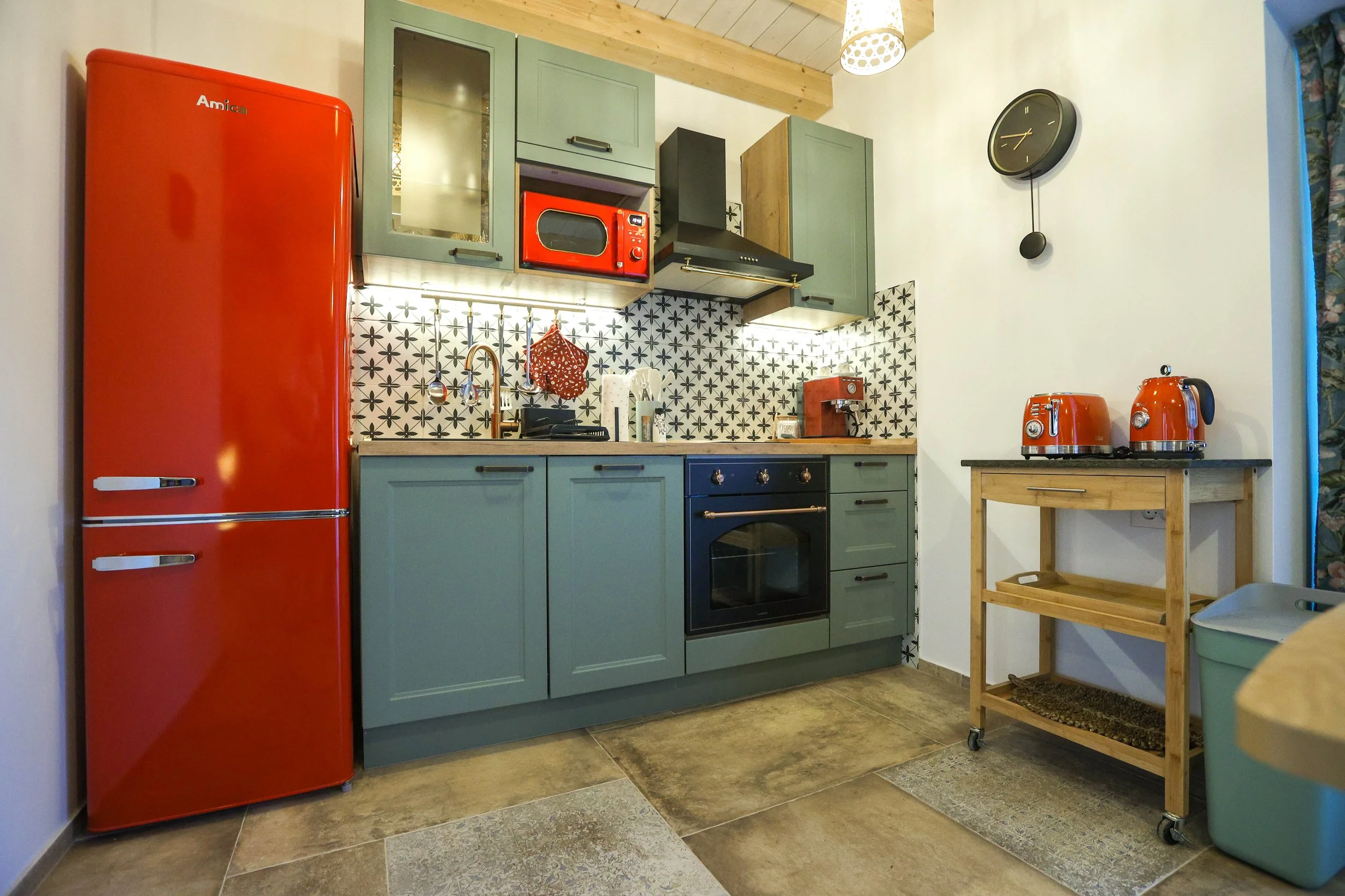A cozy kitchen with green cabinets, a red refrigerator, a black oven, and small appliances on the counter, including an orange toaster and kettle. The backsplash has a black and white star pattern, and there is a small wooden rolling cart on wheels to the right with additional kitchen items.