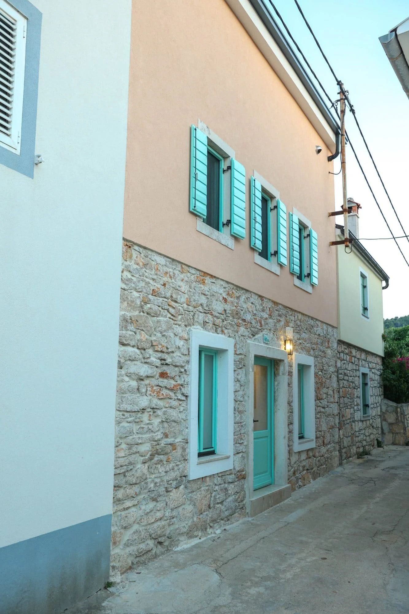 A building exterior with a stone lower half and peach-colored upper half, featuring turquoise window shutters and a door. Wires and a utility pole are visible.