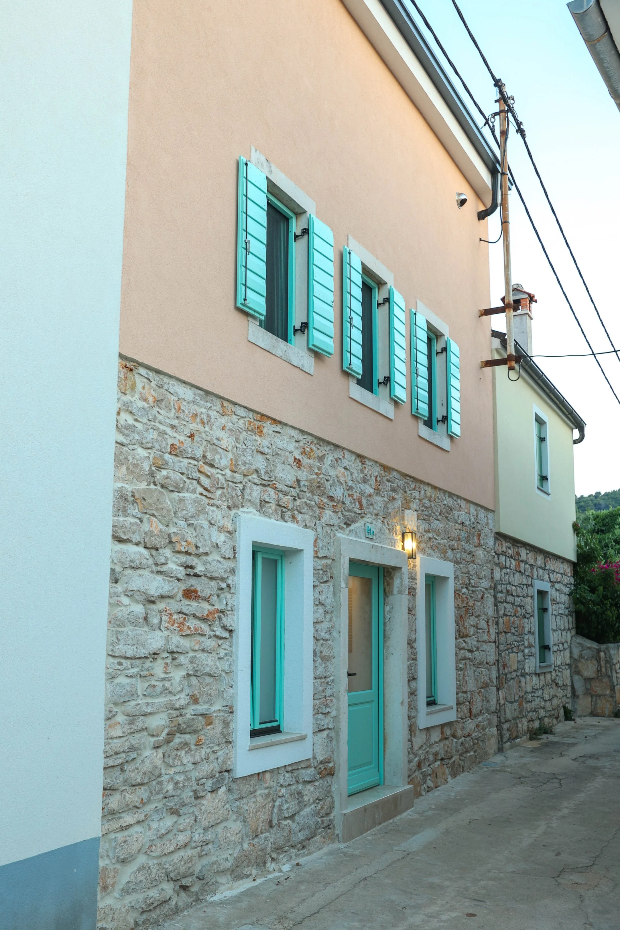 A street view of a two-story building with a stone lower level and a peach-colored upper level. The upper windows have aqua-colored shutters, and the lower windows are framed in white with aqua doors. A street lamp is mounted on the wall below the upper windows, and overhead wires run along the street.