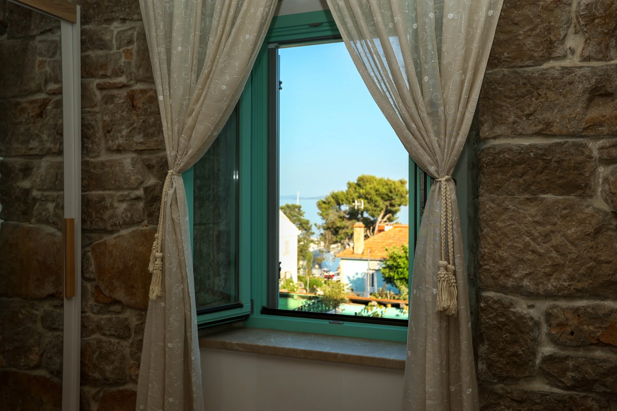 Open window with beige curtains tied back, reveals a view of a cityscape with trees, rooftops, and a body of water in the distance.