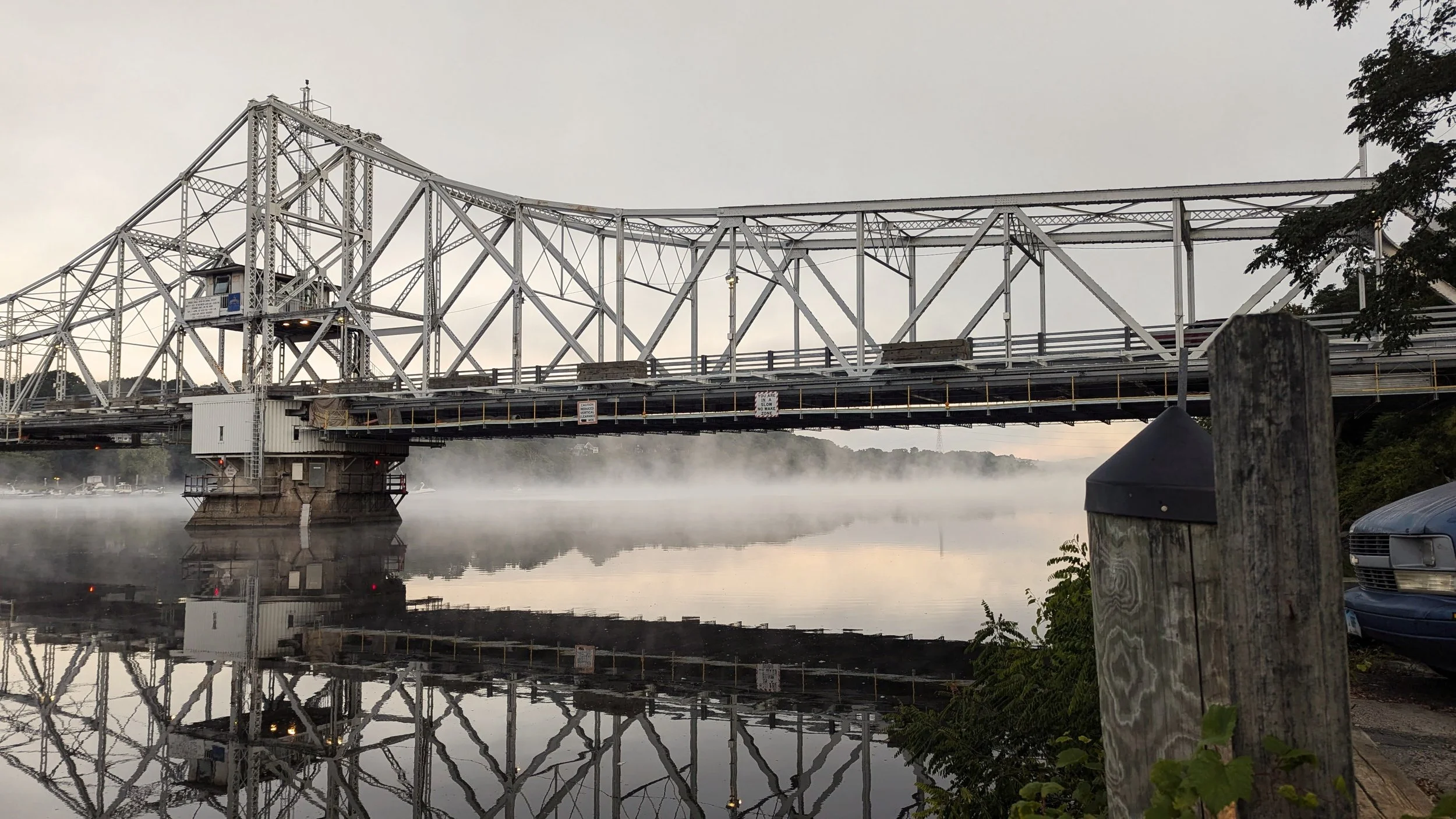 A metal truss bridge over a body of water with a misty reflection on the surface, surrounded by fog and some trees and cars visible in the background.