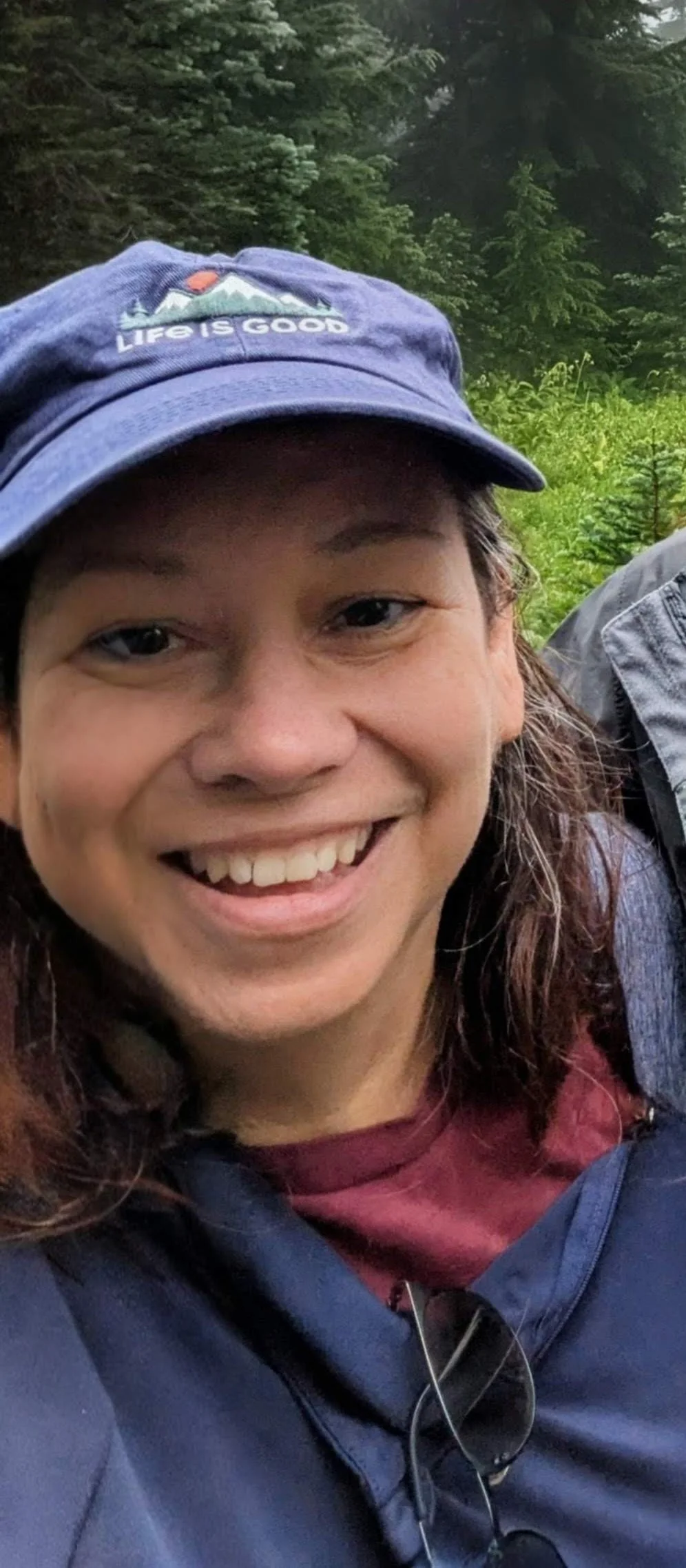 A smiling woman wearing a blue cap with mountains and the words 'Life is Good,' in a green forest.