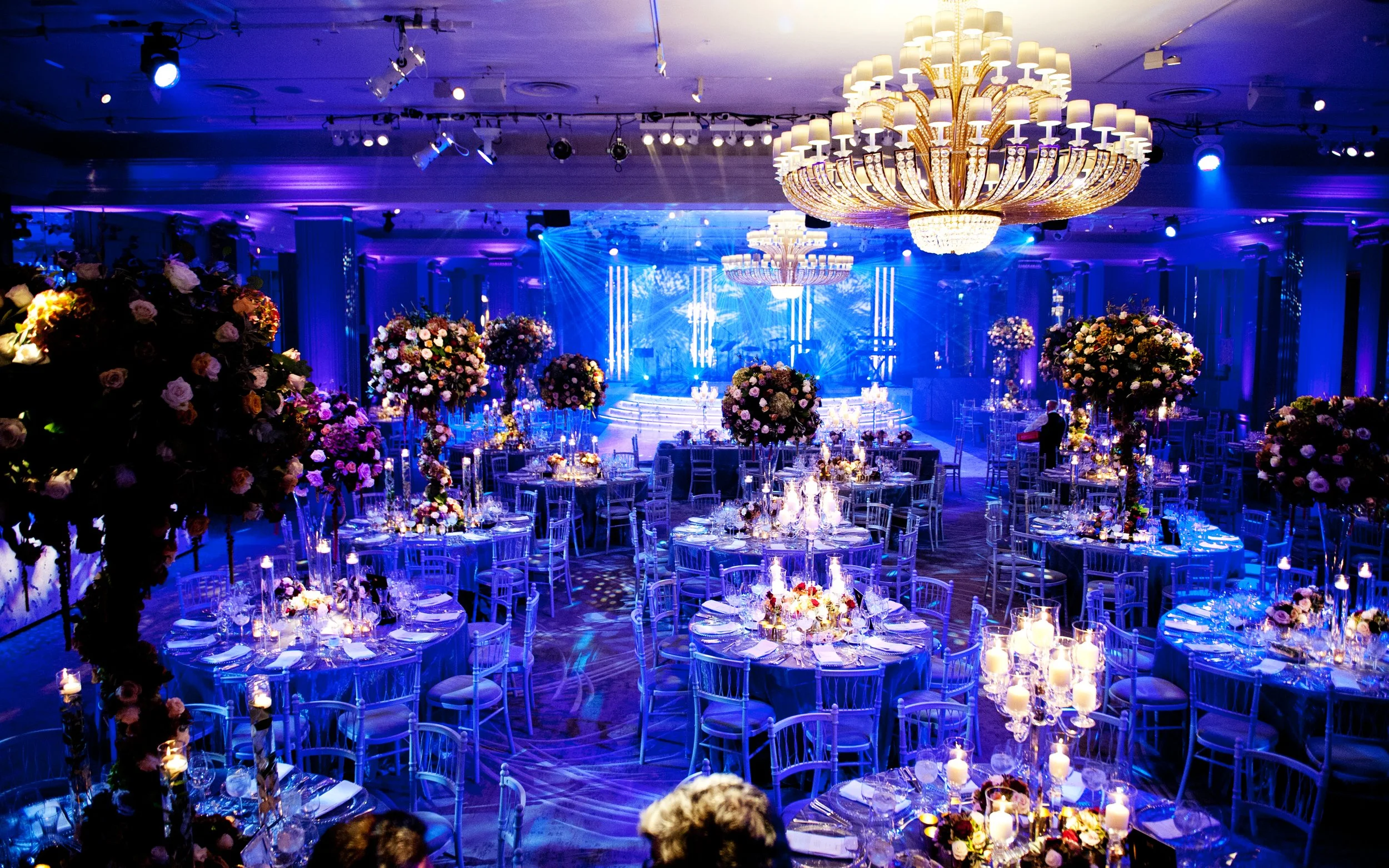 A lavish Grosvenor House for a wedding with numerous round tables, floral centerpieces, tall candles, and elegant chairs. Large chandeliers hang from the ceiling, and blue and purple lighting creates a glamorous atmosphere. 