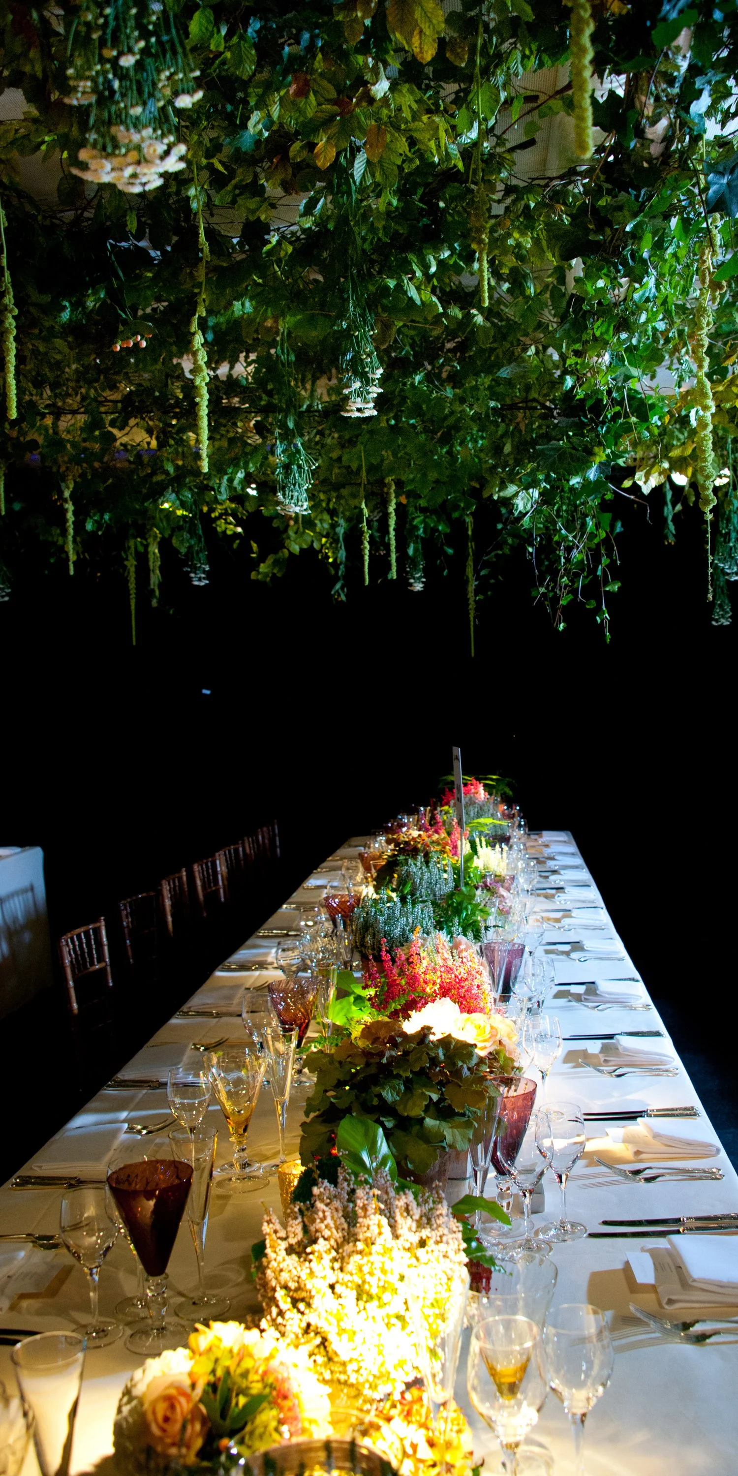 Elegant banquet table decorated with colorful flowers, glassware, and cutlery, set beneath a lush, green leafy overhead installation for an event or celebration.