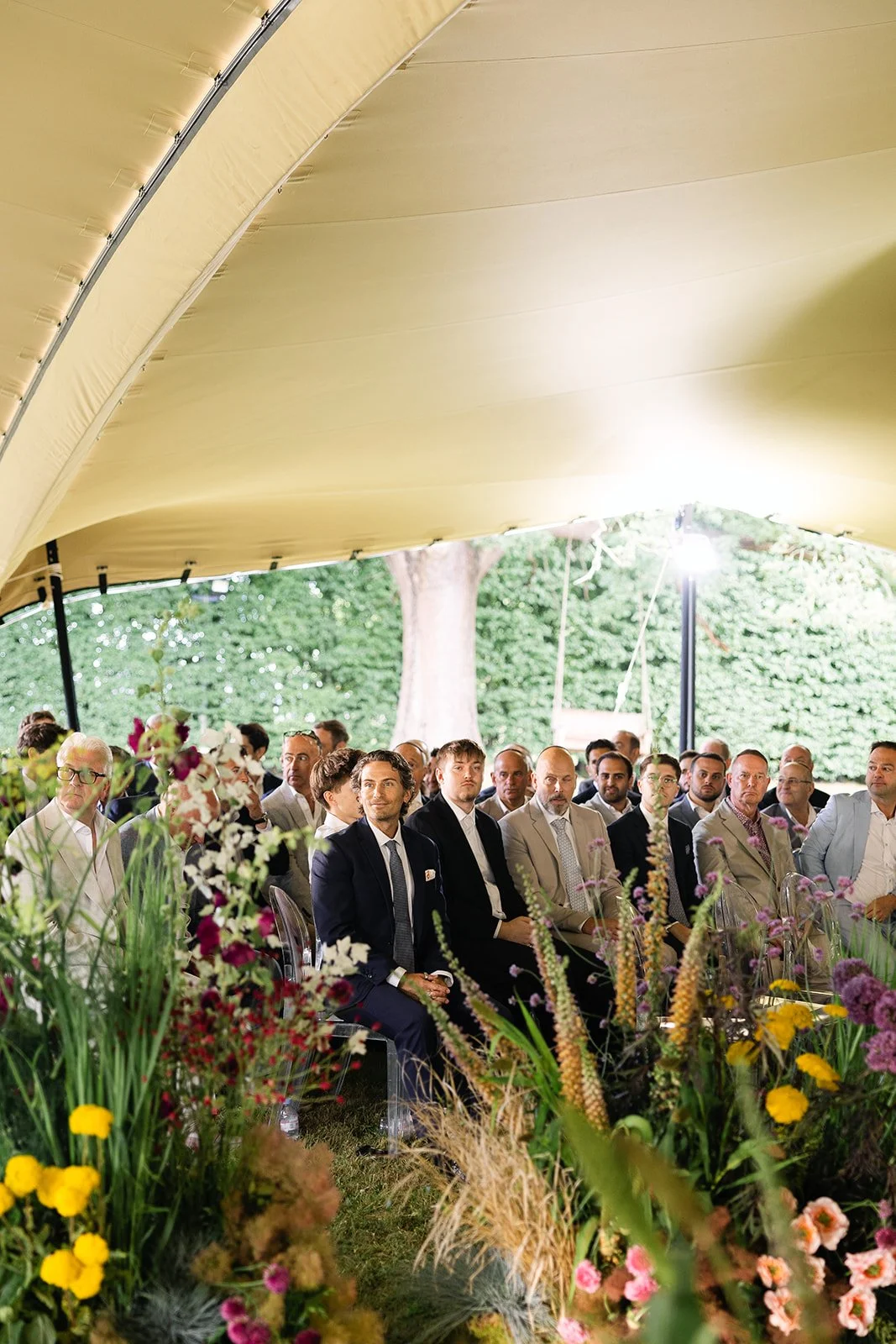 Wedding guests at a jewish wedding under a beige canopy.  The venue has lush greenery and a large tree visible in the background. Colorful flowers are arranged in the foreground.