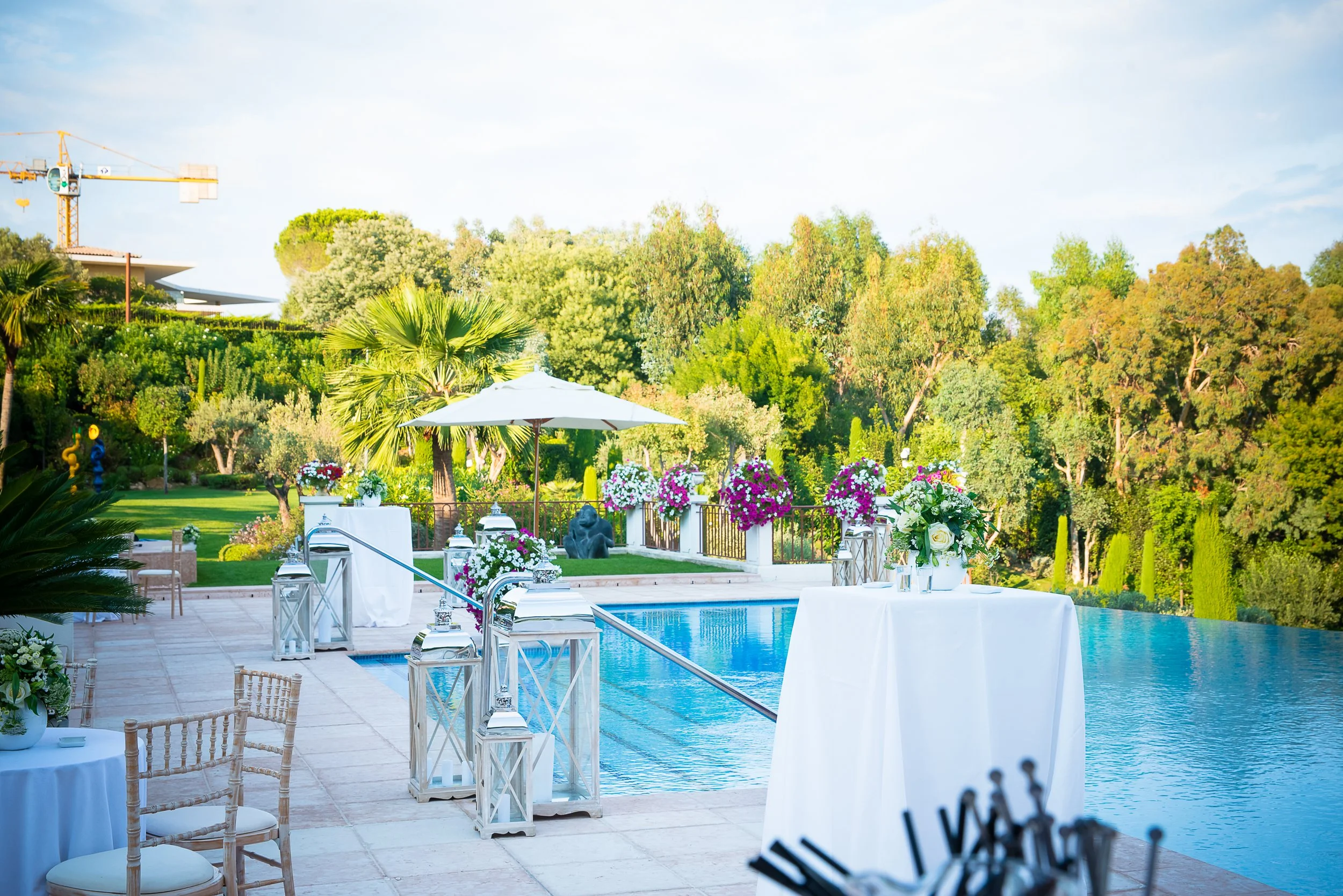 Elegant outdoor poolside area set up with white tables and chairs, floral arrangements, and lanterns, with a backdrop of lush green trees and a clear sky, suggesting a setting for a wedding or special event.