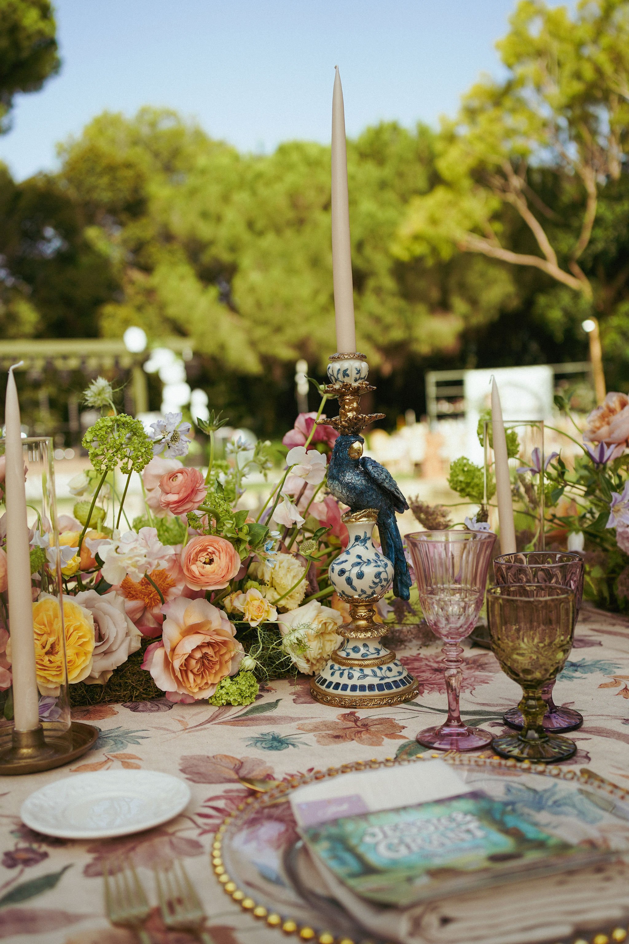 Table decorated with a floral tablecloth, pink, yellow, and purple flowers, and a blue and white ornate candlestick with a tapered candle, surrounded by pink and green glassware glasses.