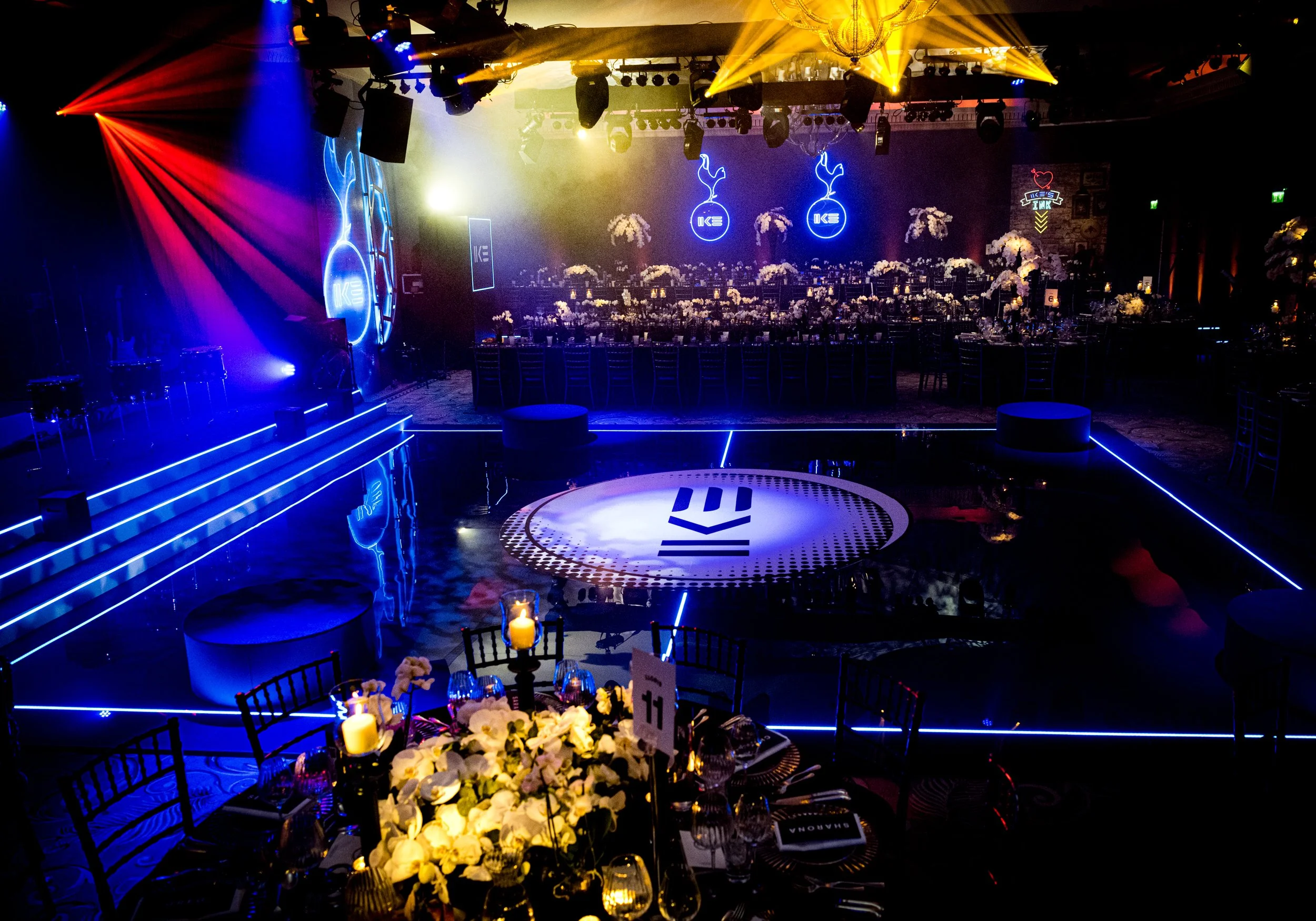 A dimly lit event space with colorful stage lighting, floral arrangements, and tables set for a formal event for a Football themed luxury Bar Mitzvah