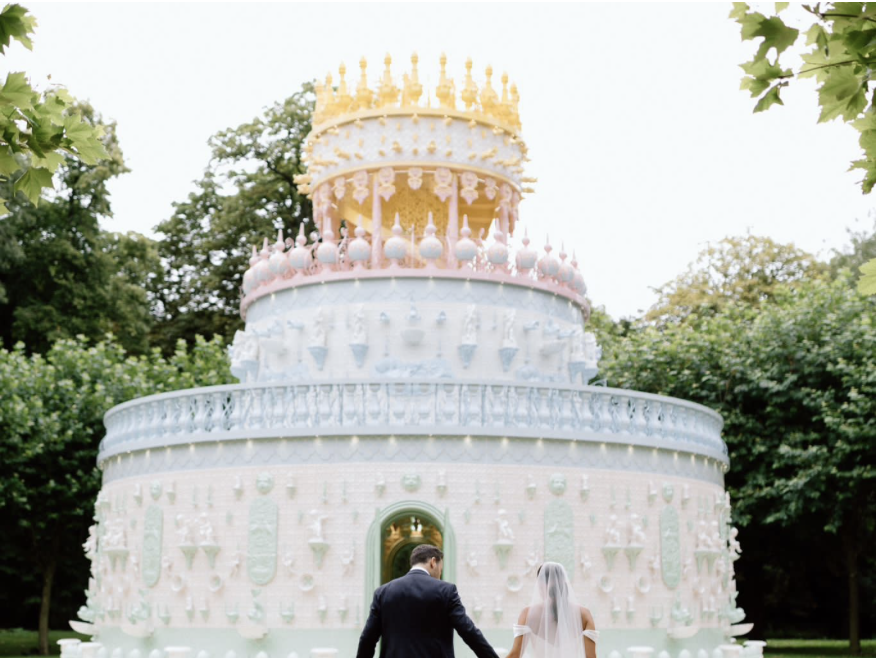 A couple in wedding attire holding hands in front of a large, colorful tiered cake-shaped sculpture outdoors surrounded by green trees.