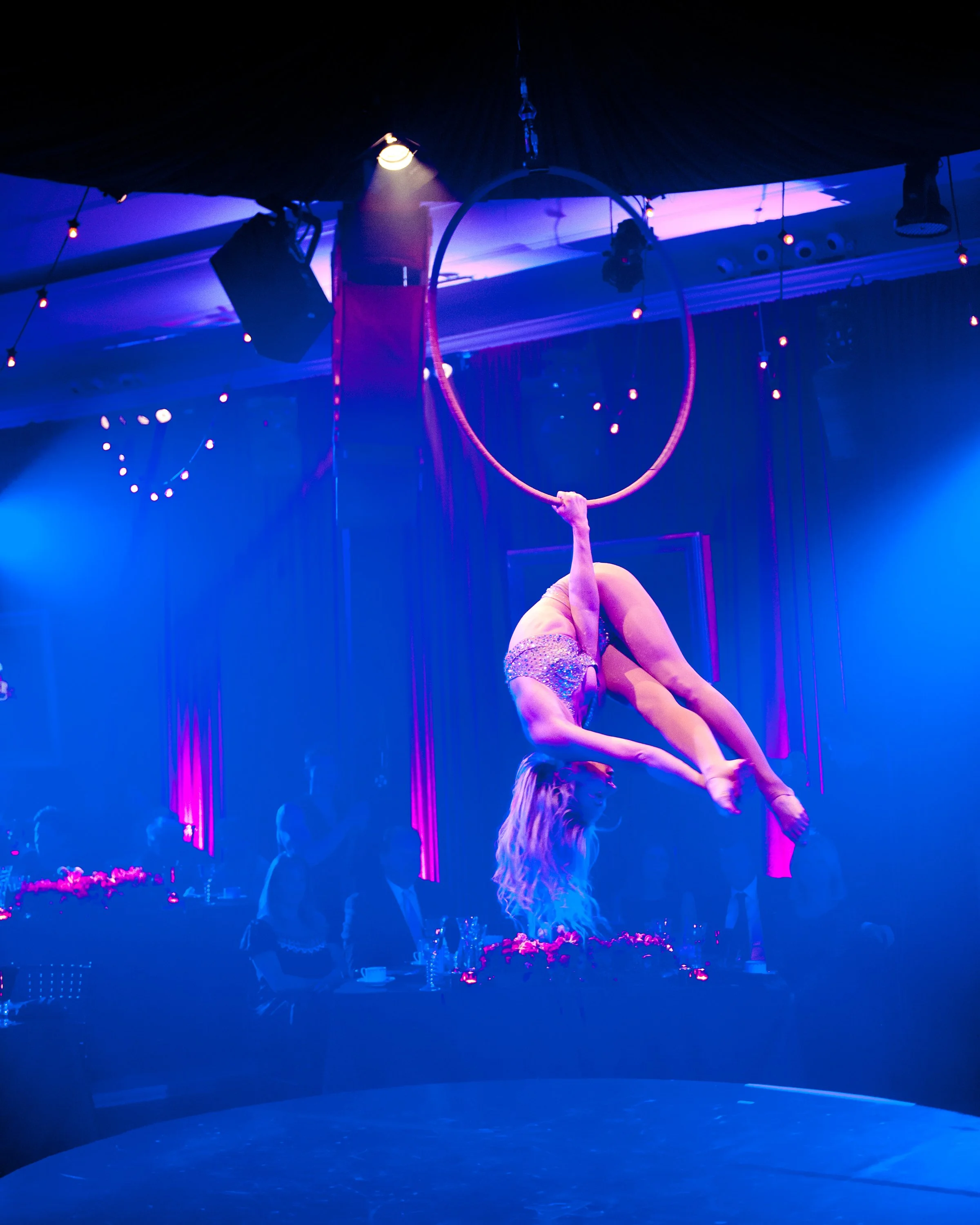 A performer is suspended upside down on a hoop, decorated with sparkling rhinestones, in a dark, blue-lit circus or acrobatic show.