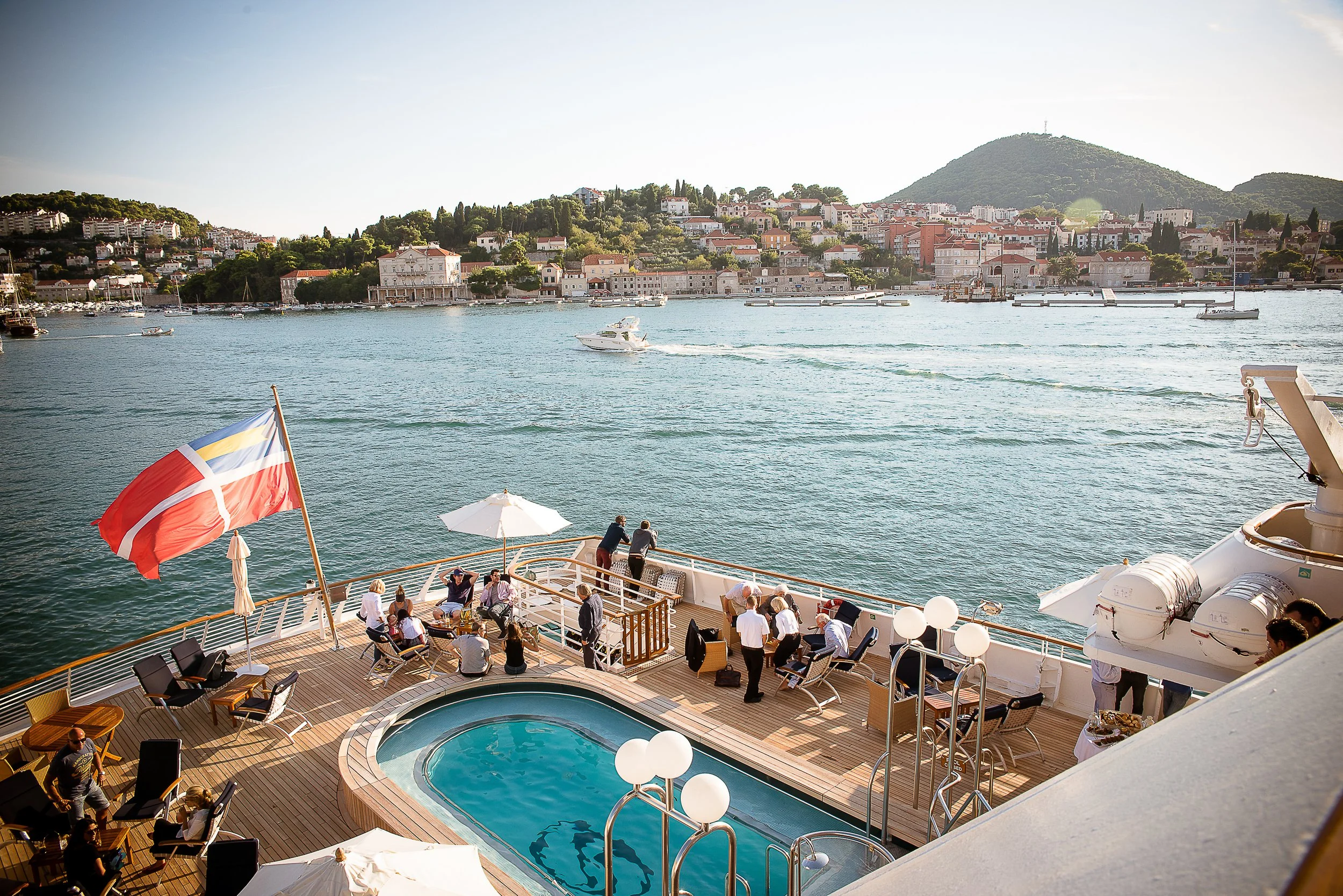 A view from a cruise ship deck showing an outdoor pool surrounded by seating and tables, with people relaxing and socializing. The ship is floating on water with boats and yachts nearby. In the background, there is a coastal town with white buildings, red-tiled roofs, and green hills.