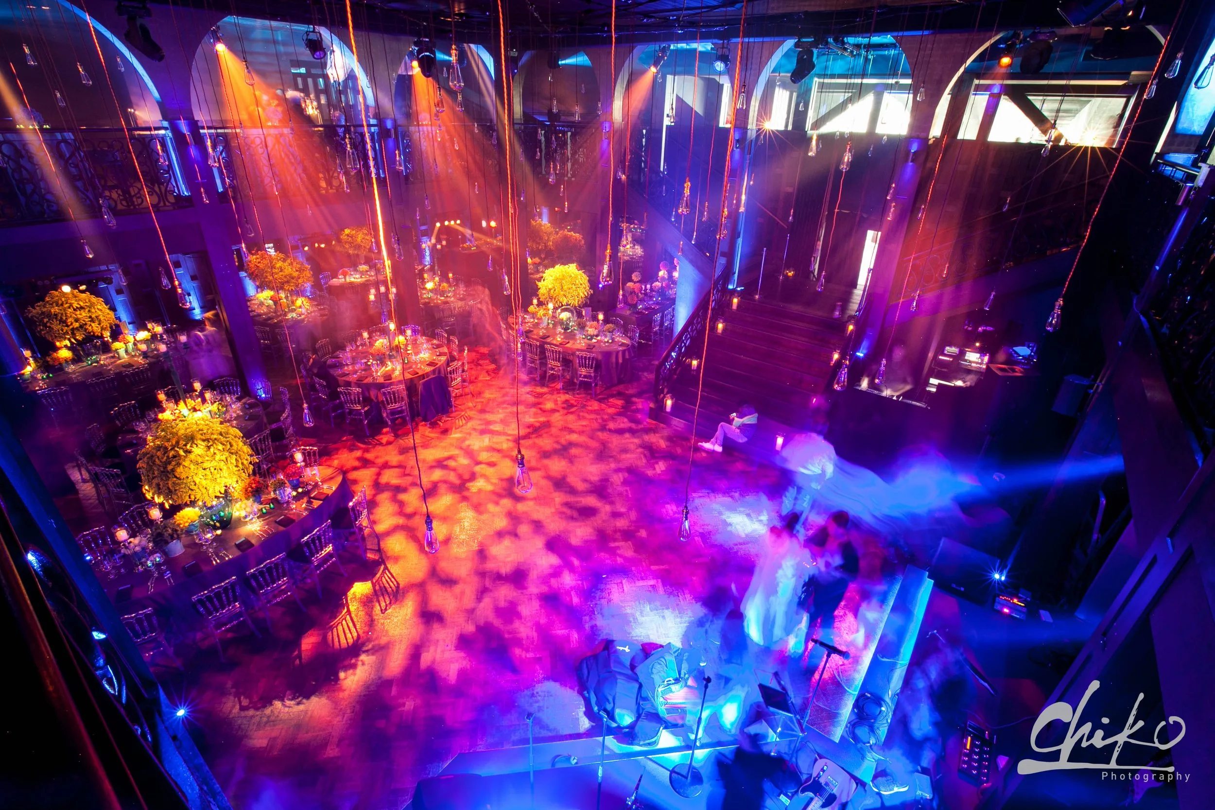 A dimly lit event space with colorful blue, purple, and orange lighting, decorated with hanging Edison bulbs and floral centerpieces on round tables surrounded by chairs. There is a balcony railing on the left side and stairs leading up on the right 