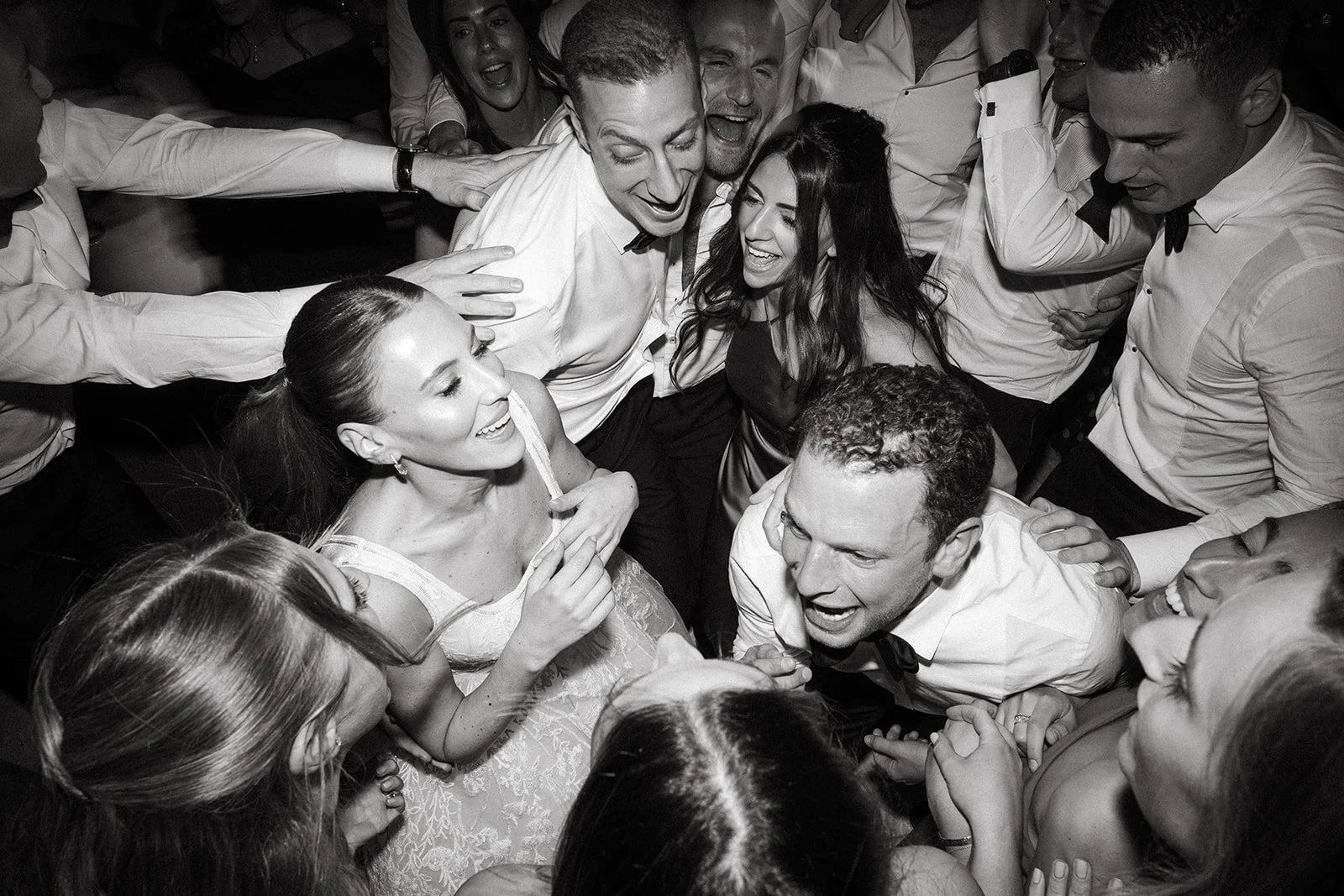 Black and white photo of a group of people, all dressed formally, gathered closely and excited, sharing a joyful moment, with smiles and laughter at The Peninsula London. Wedding party