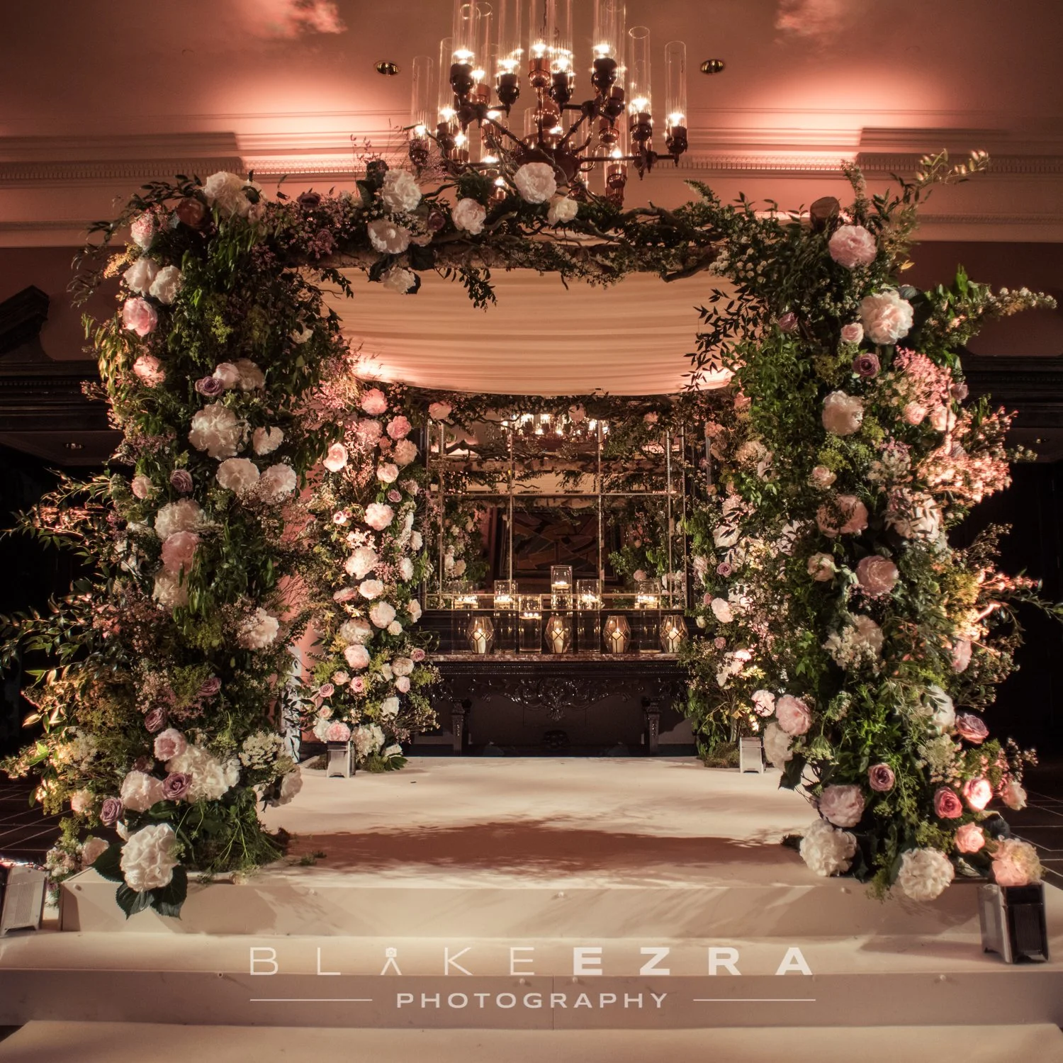 Elegant wedding arch decorated with pink and white flowers, greenery, and a draped fabric canopy, with a chandelier hanging above and candles on a table in the background.