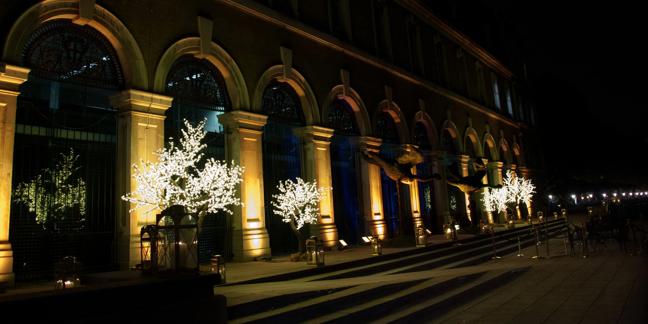 Nighttime view of a building facade with arched windows and columns illuminated by warm lights, decorated with white string lights on small trees and topiary in front, along with lanterns lining the steps leading up to the entrance.