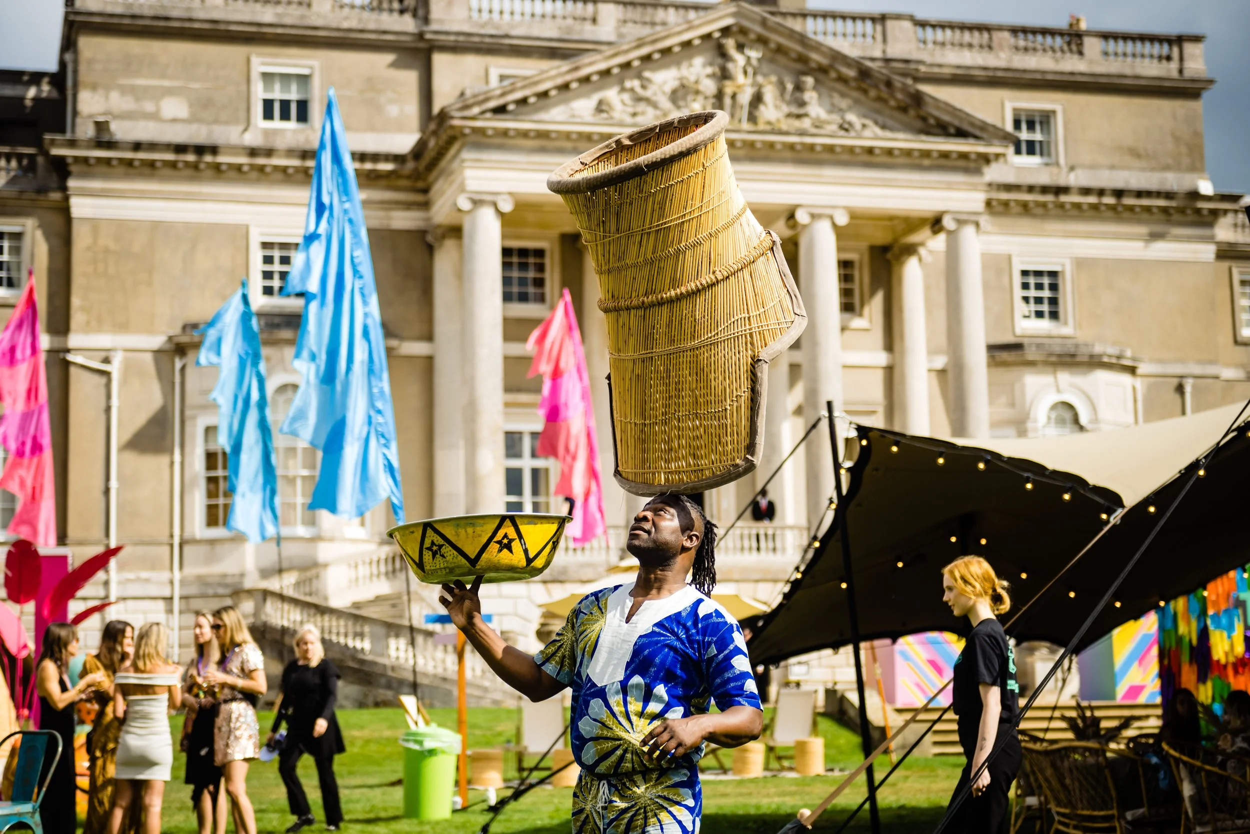 A man with comically large balancing bowls on his head, performing at an outdoor festival or parade with colorful flags and crowd in the background, in front of a historic building at a Bar Mitzvah at  Wrotham Park