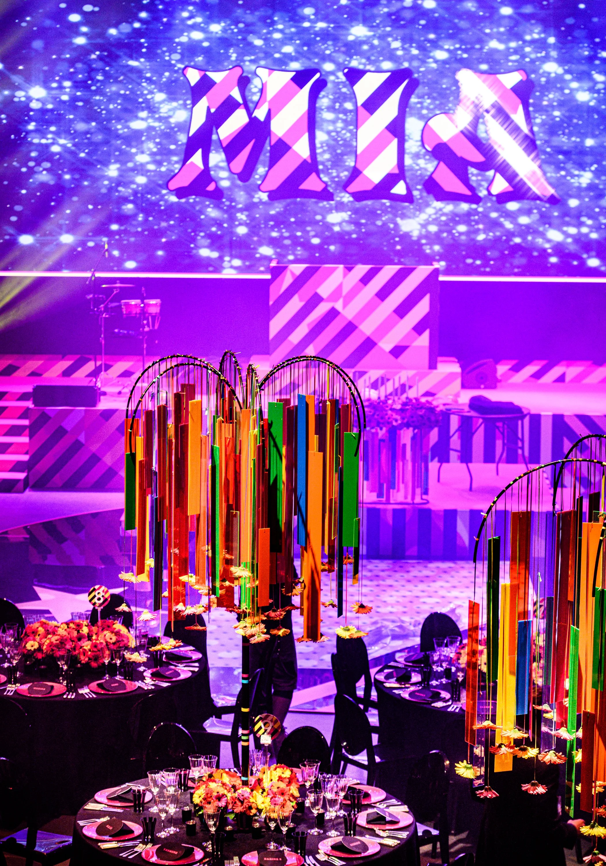 Decorated event space with purple lighting, large colorful hanging art installations, tables set with floral centerpieces, black tablecloths, and place settings for a formal gathering.  Bat Mizvah the roundhouse