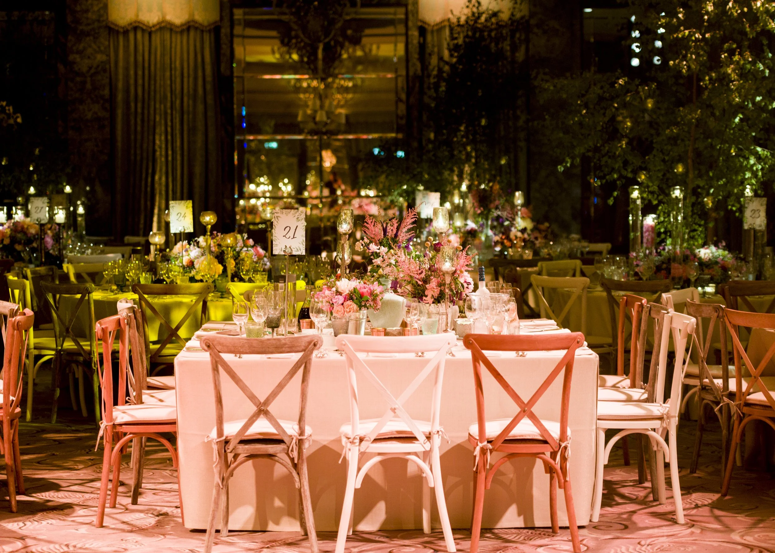 Elegant dining setup with arranged tables, pink flowers, and candles in a warmly lit room decorated for an event.