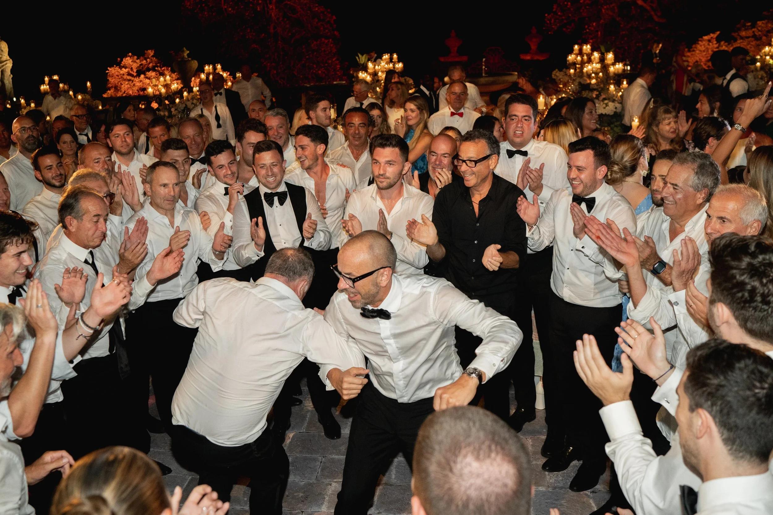 A large group of people dressed in formal attire at a celebration, with many clapping and smiling, in a decorated outdoor setting at night at Villa Palmieri in Florence