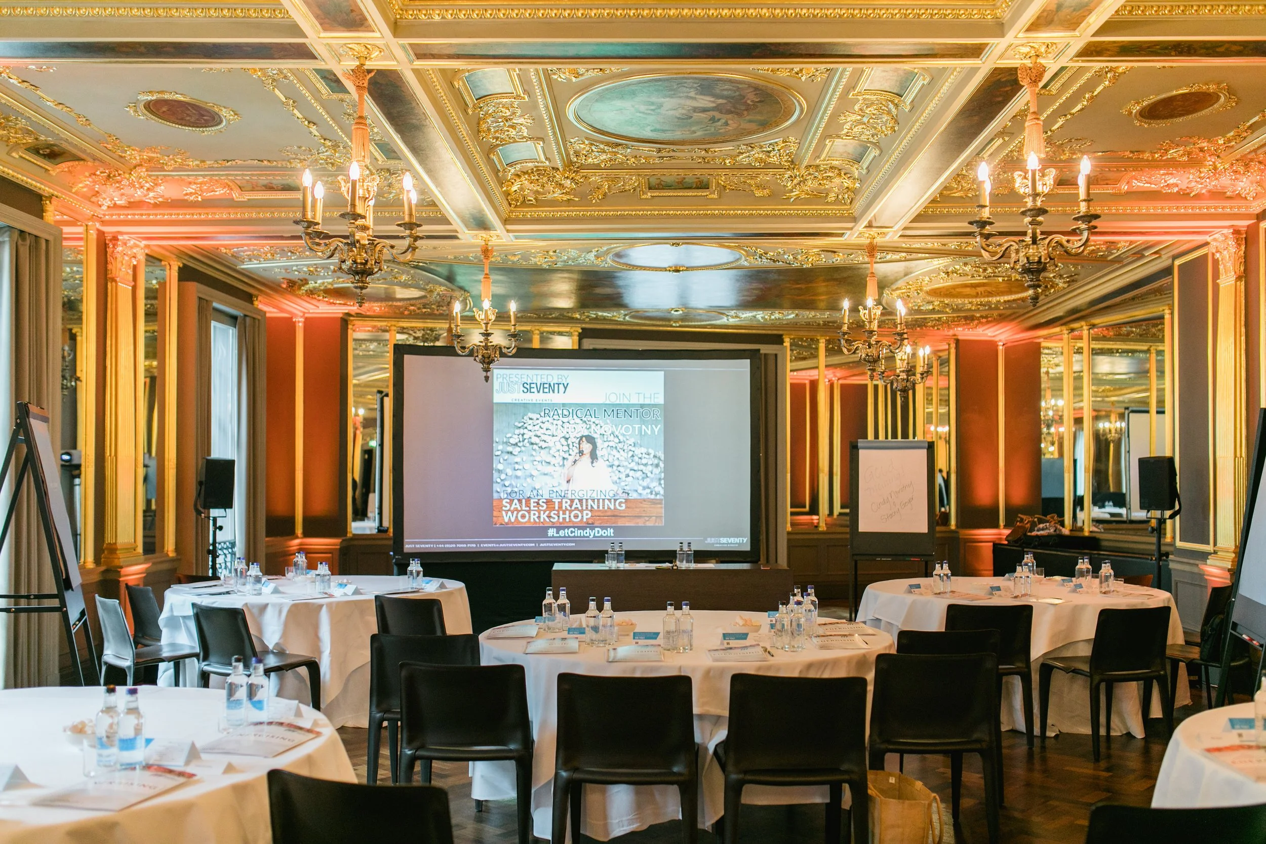 A conference room with ornate gold and pastel ceiling, chandeliers, and round tables covered with white tablecloths, set with water bottles, papers, and notepads, with a large screen displaying a presentation at the front.