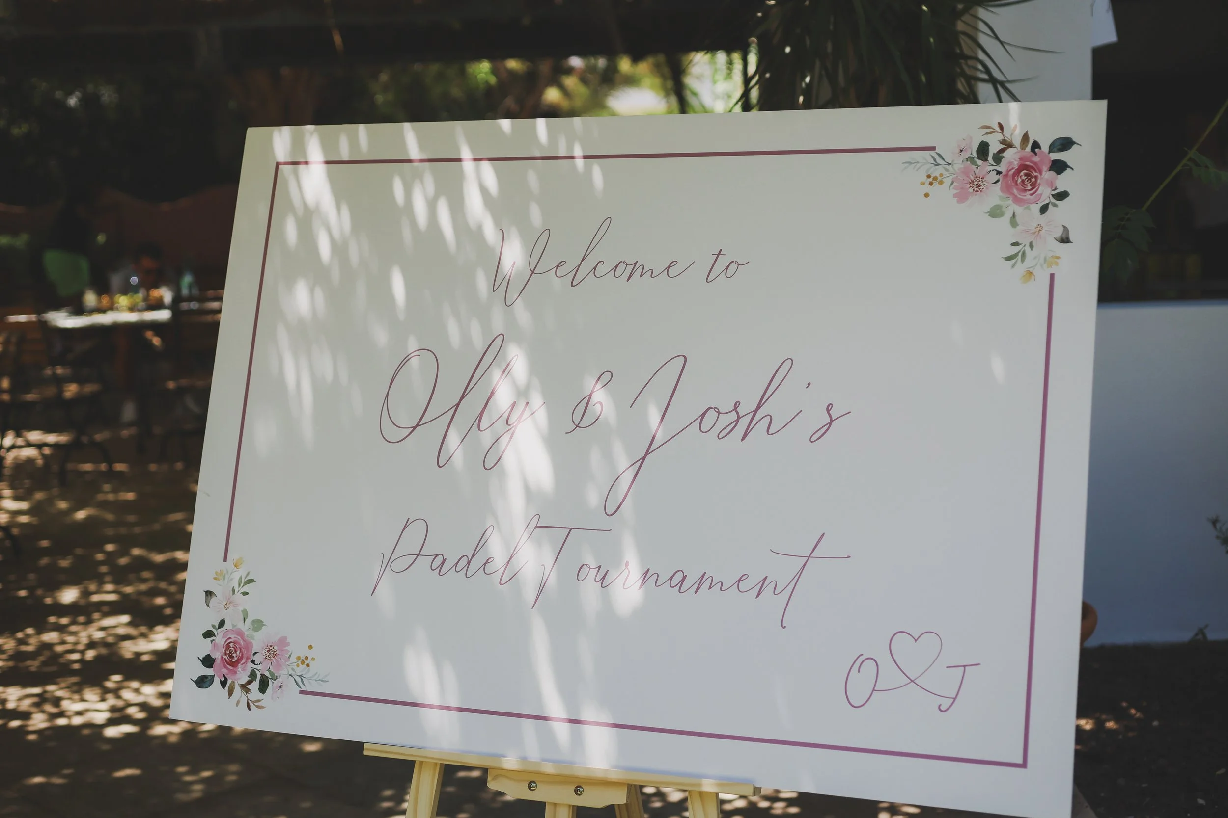 A welcome sign with floral decorations at a wedding event. It reads 'Welcome to Ely & Josh's' and mentions a 'Paddle Tournament,' with some of the text partially obscured or out of frame.