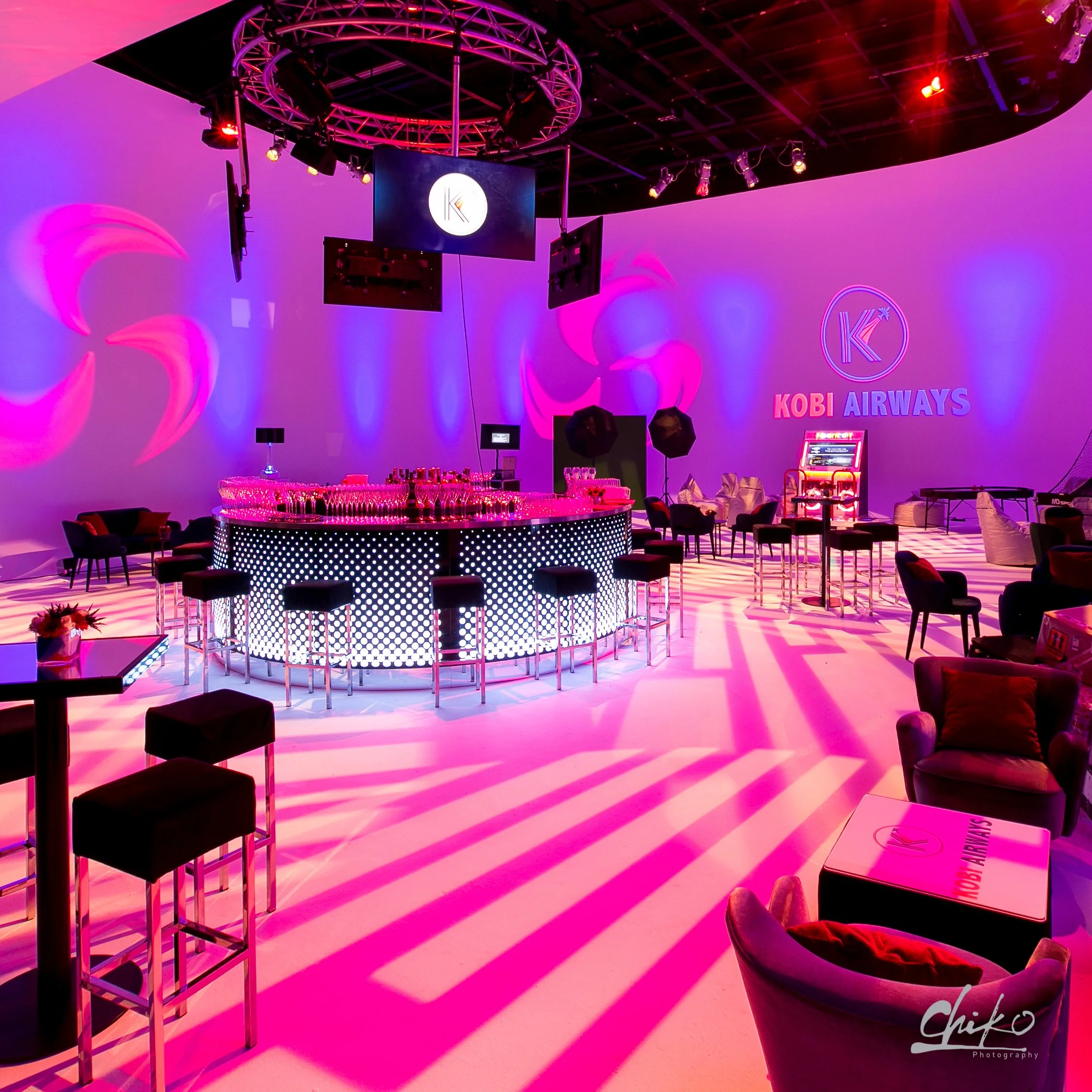 Interior of a nightclub or party venue with a bar illuminated by pink and purple lighting. The bar is round with a dotted light pattern and drinks arranged on top. Surrounding the bar are black chairs and tables, with some decorated with flowers. The