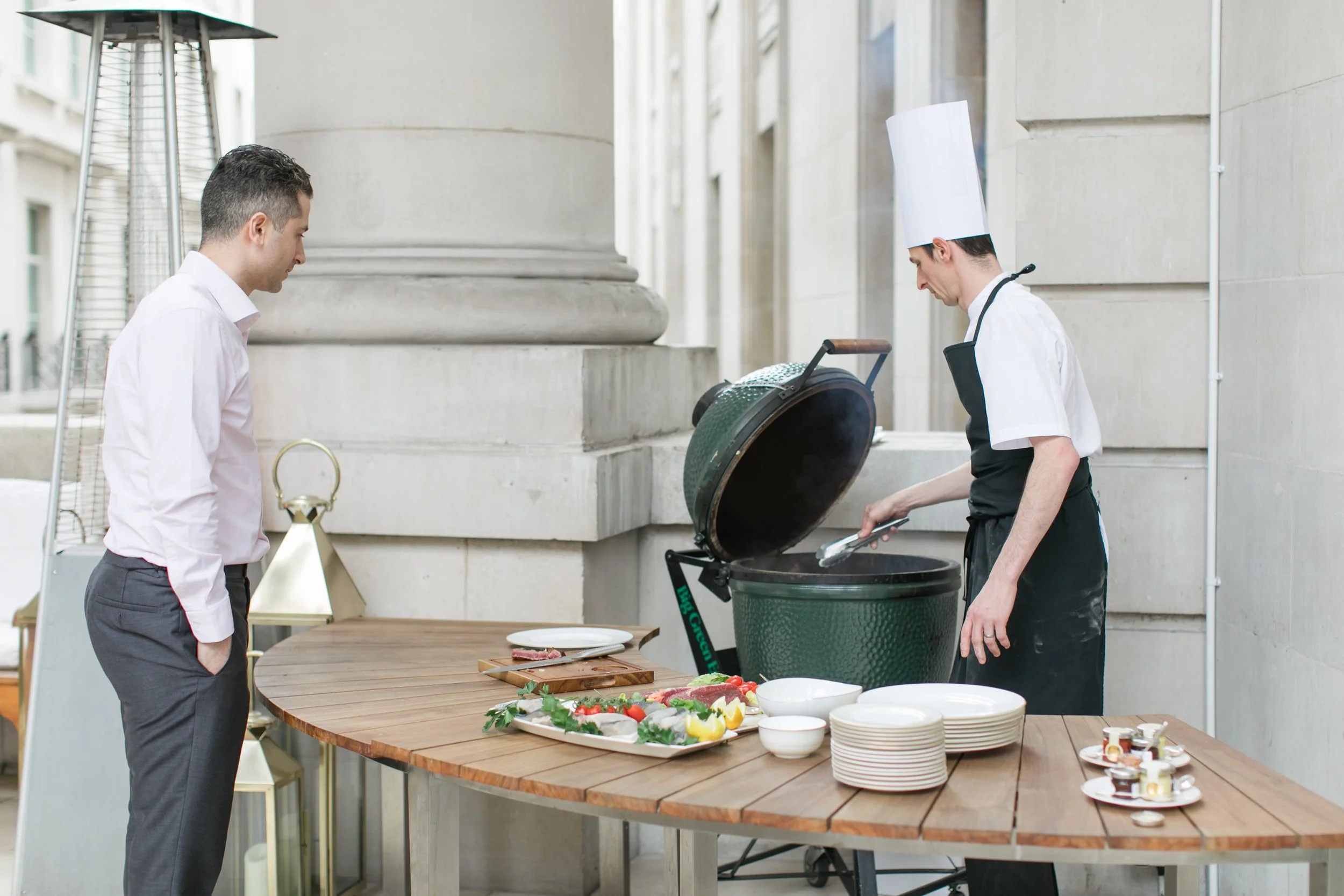 A man in a white shirt and gray pants watches a chef in a white uniform and tall chef's hat as the chef grills food outdoors. There is a round wooden table with plates of sliced vegetables, fresh herbs, and various condiments in front of them.