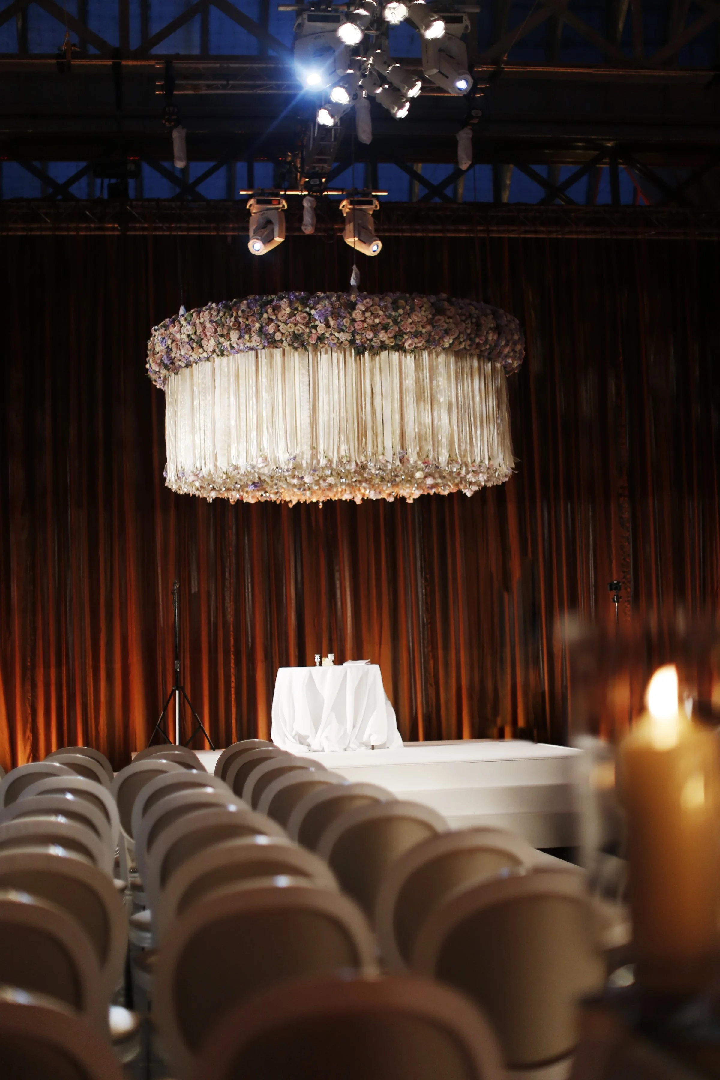 Empty chairs facing a stage with a table covered in a white cloth, and a large floral chandelier hanging from the ceiling, with brown curtains in the background.