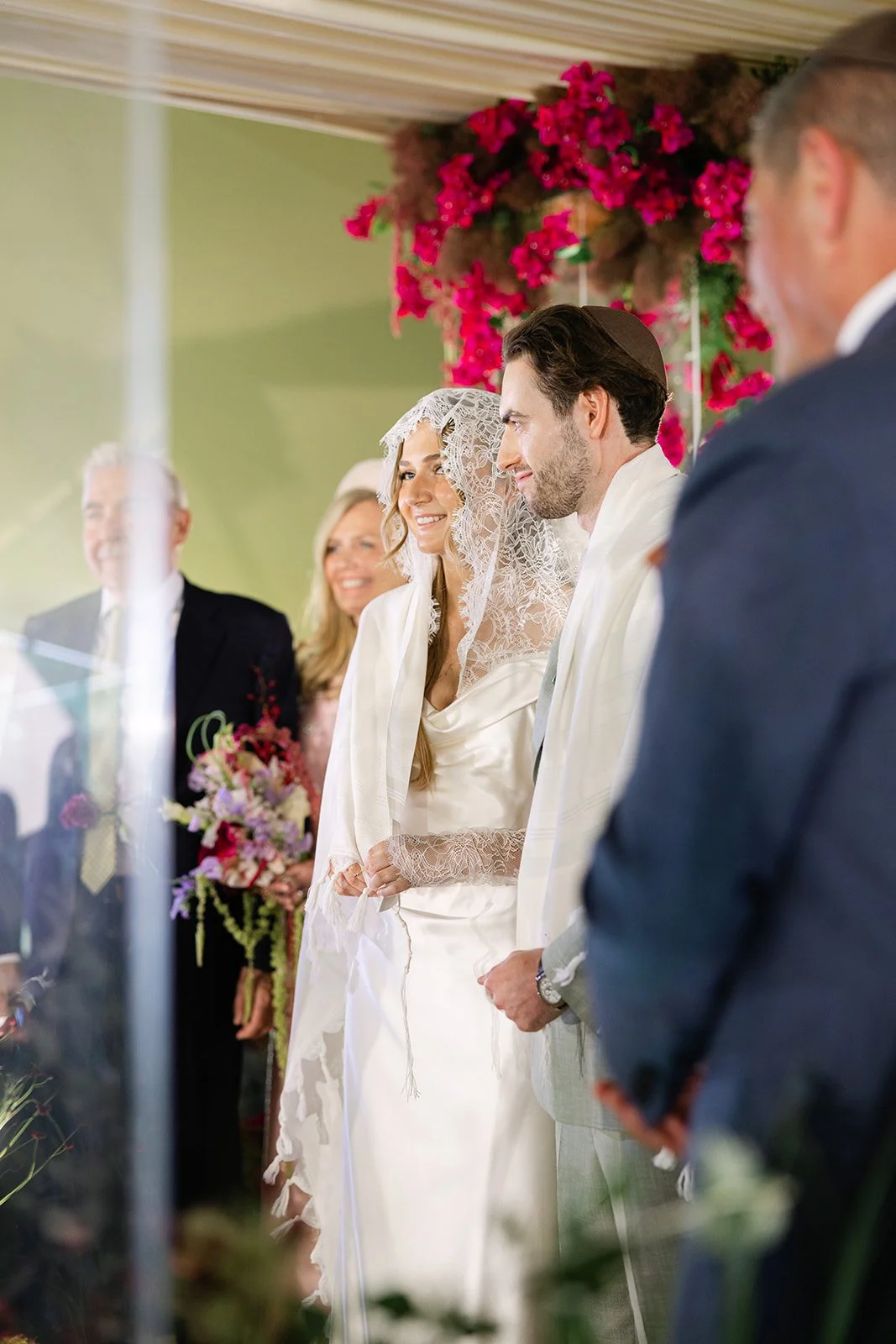 A wedding ceremony a decorated chuppah and venue with pink flowers in the background. The bride, wearing a lace veil and satin gown, stands with the groom in a light suit