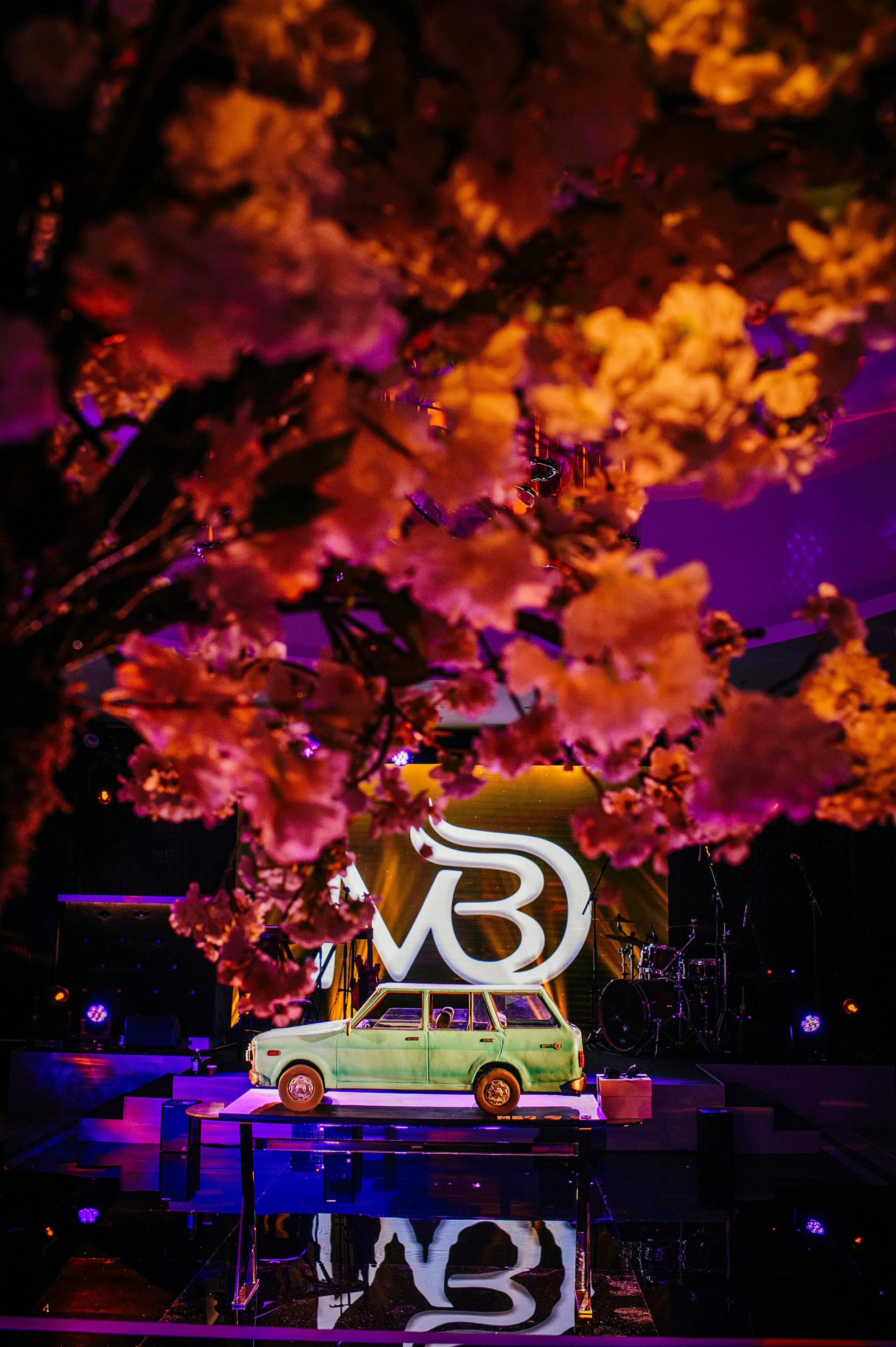 Stage setup with a vintage green car, large illuminated logo with the letter 'B', pink cherry blossom tree, and musical instruments in the background, illuminated by purple and orange lighting.
