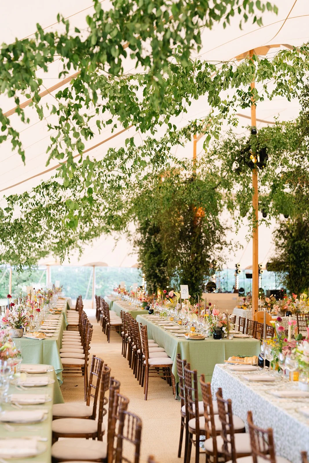 A decorated indoor event space with tables set for a formal dinner, covered with green tablecloths and adorned with pink and white floral centerpieces, underneath a ceiling draped with green leafy foliage.