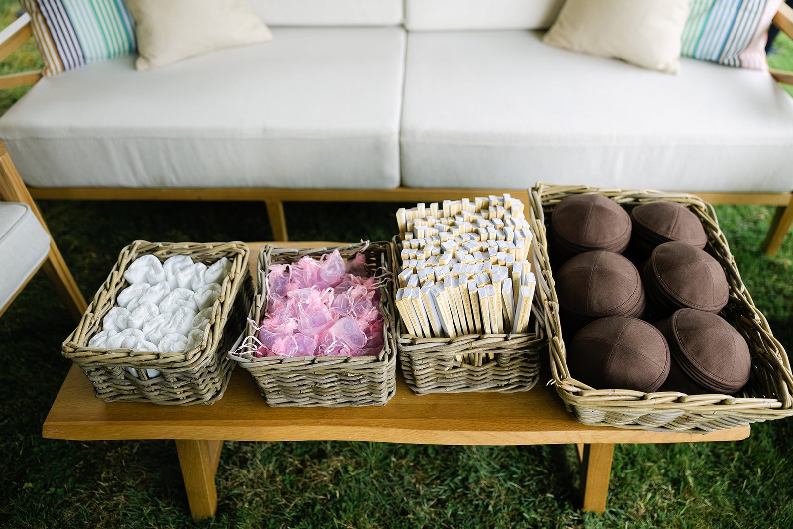 A wooden table with baskets of white, pink, and brown objects such as fans, kippahs in front of a white outdoor sofa with striped and cream pillows.