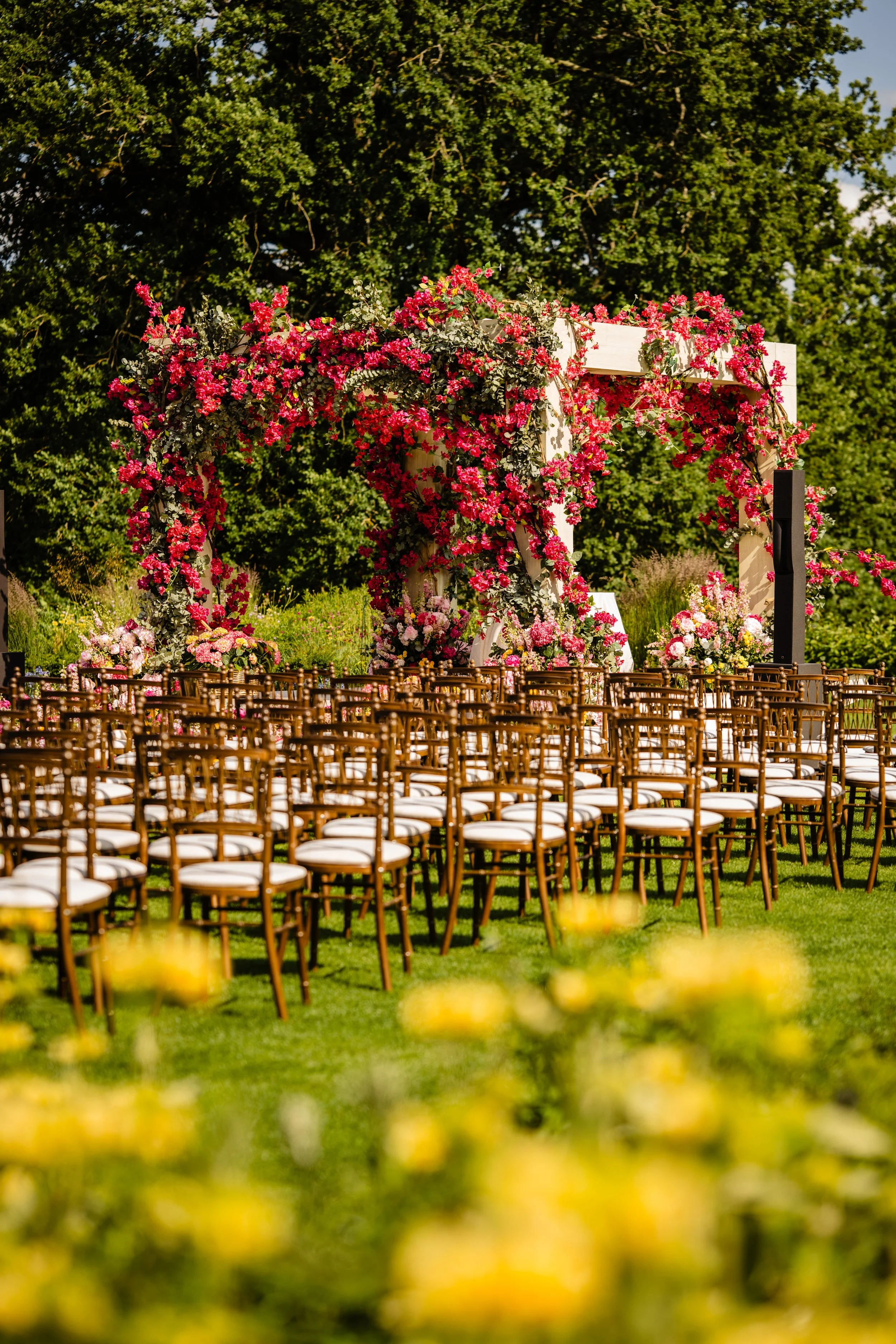Outdoor wedding setup with rows of chairs facing a floral arch on a grassy lawn, surrounded by trees and a blue sky. Fairmont Windsor Park