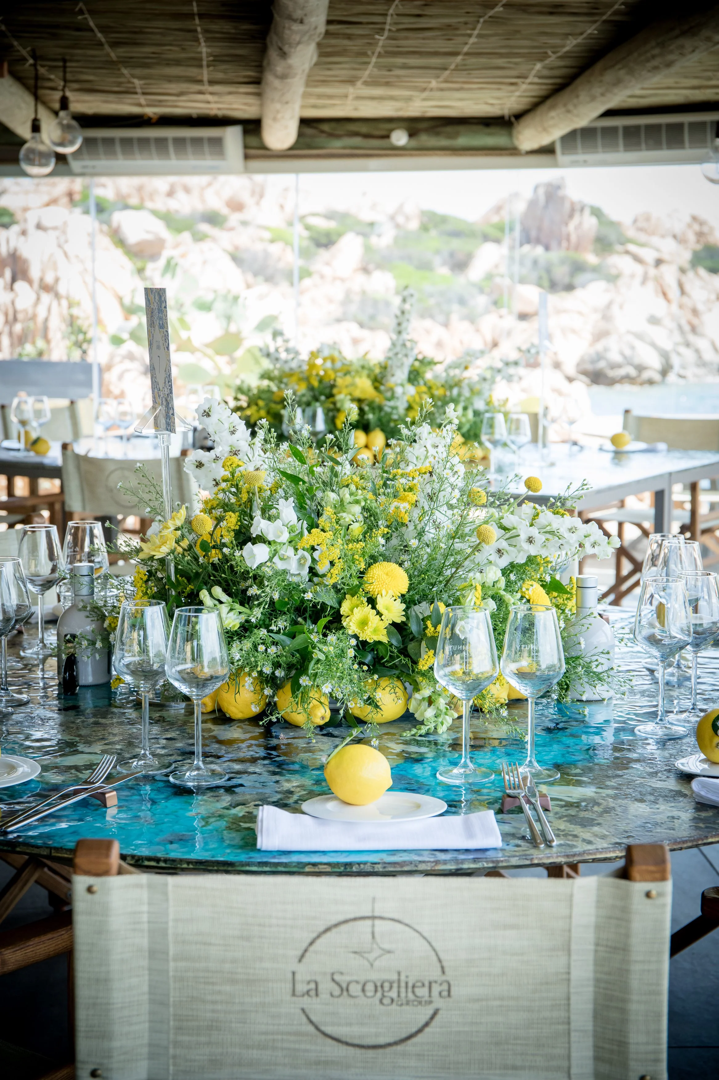 A table set for a meal featuring a large floral centerpiece with white and yellow flowers and lemons. The table has multiple wine glasses and place settings. In the background, outdoor scenery with rocks and greenery is visible through large windows,
