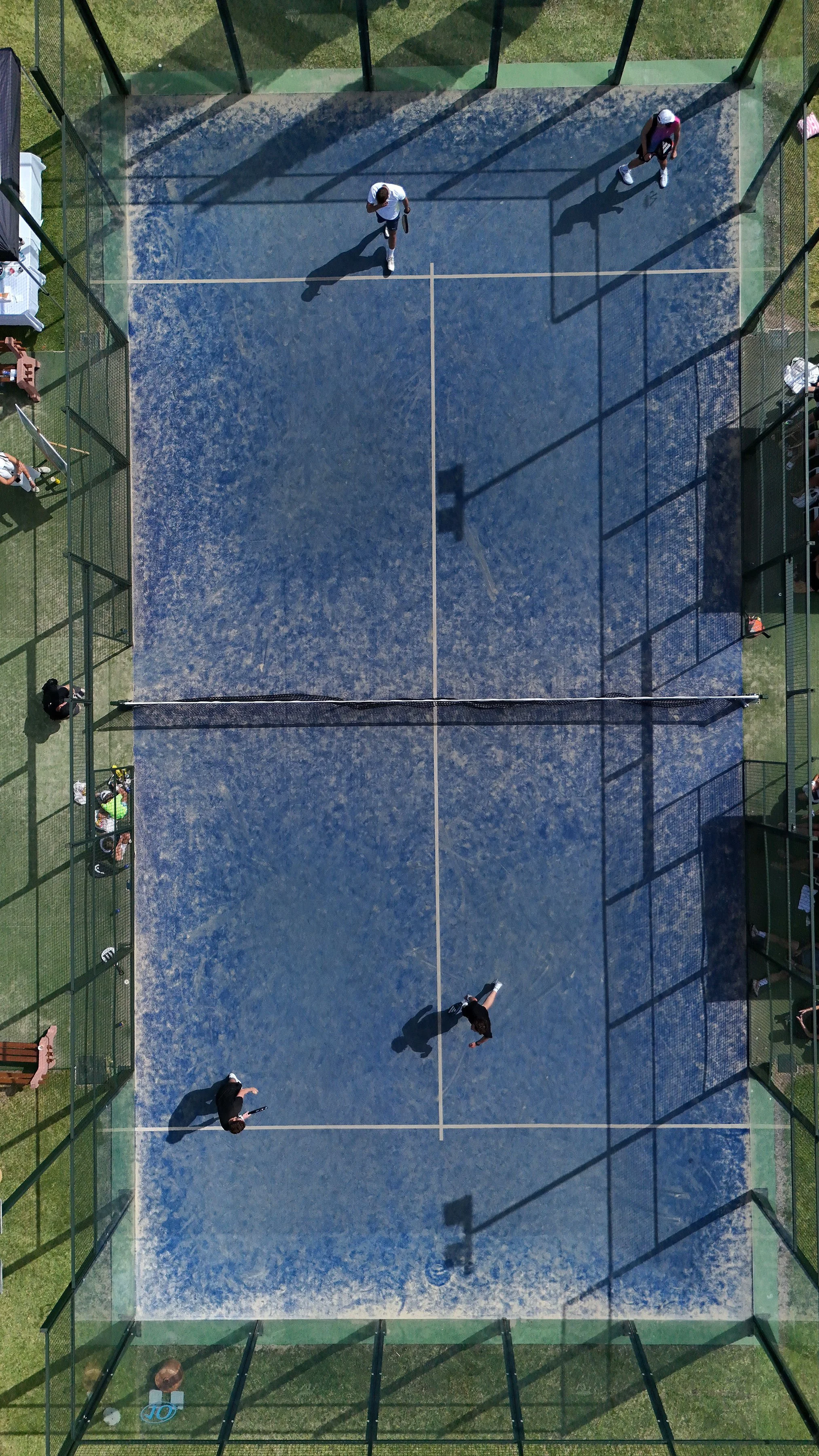 An aerial view of a paddle tennis court with four players, two on each side, and spectators on the sides. The court is enclosed with glass walls and cast shadows from the fence and players at Anantara Villa Padierna Resort in Marbella