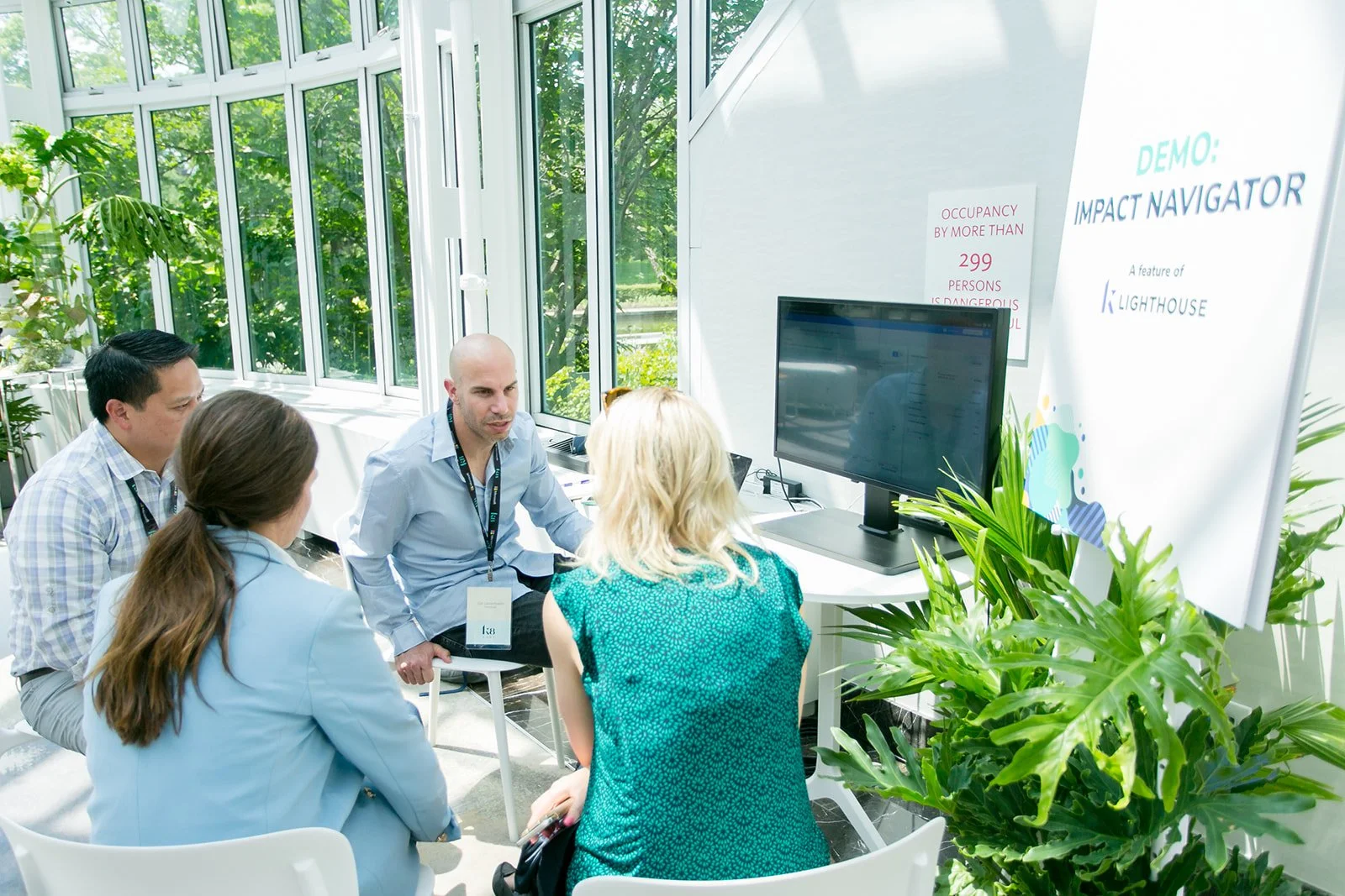 People attending a presentation or discussion in a well-lit room with large windows, green plants, and a large monitor displaying a screen, as the presenter talks to the group.